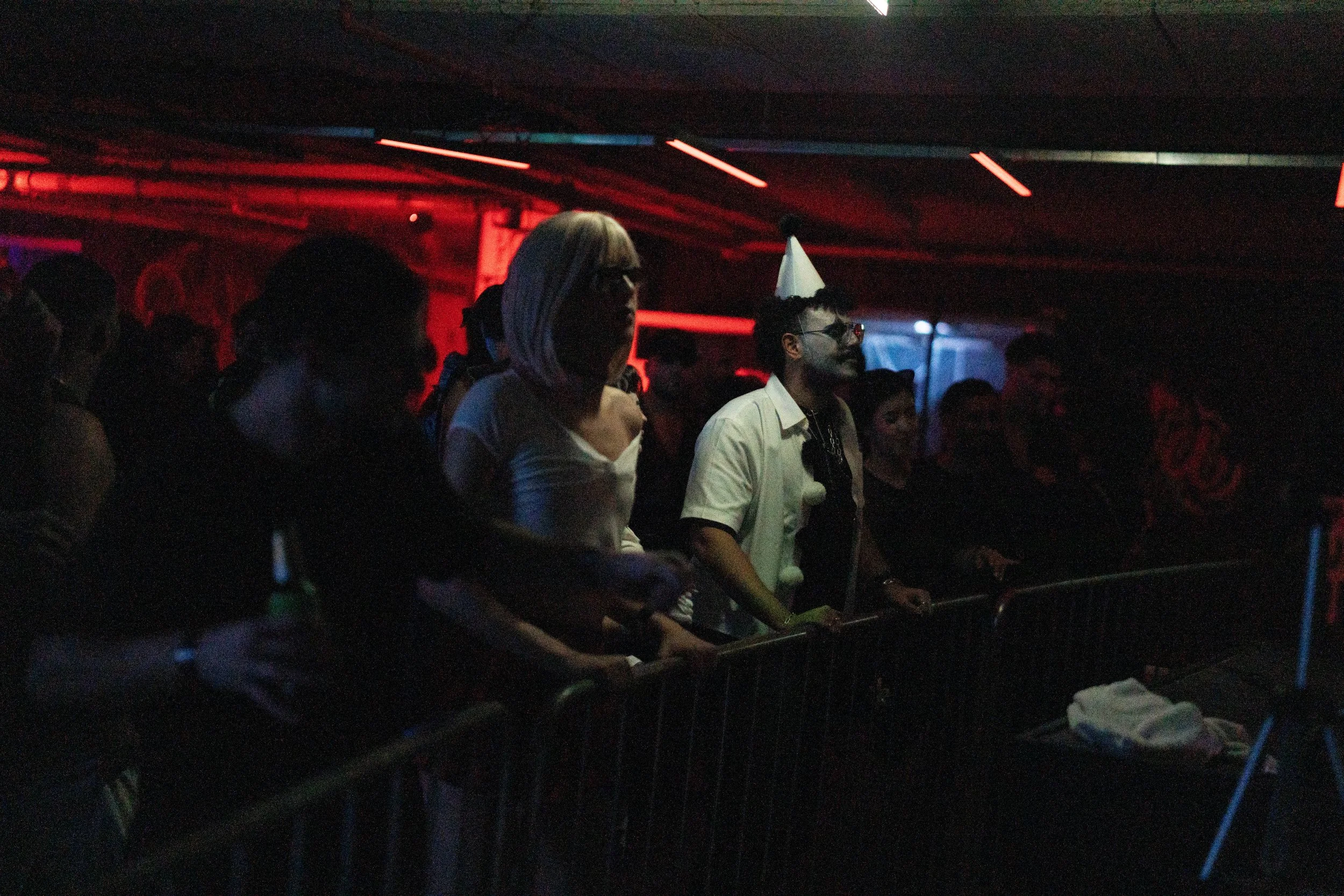 People at a party or event, some wearing party hats, in a dimly lit room with red and blue lighting.