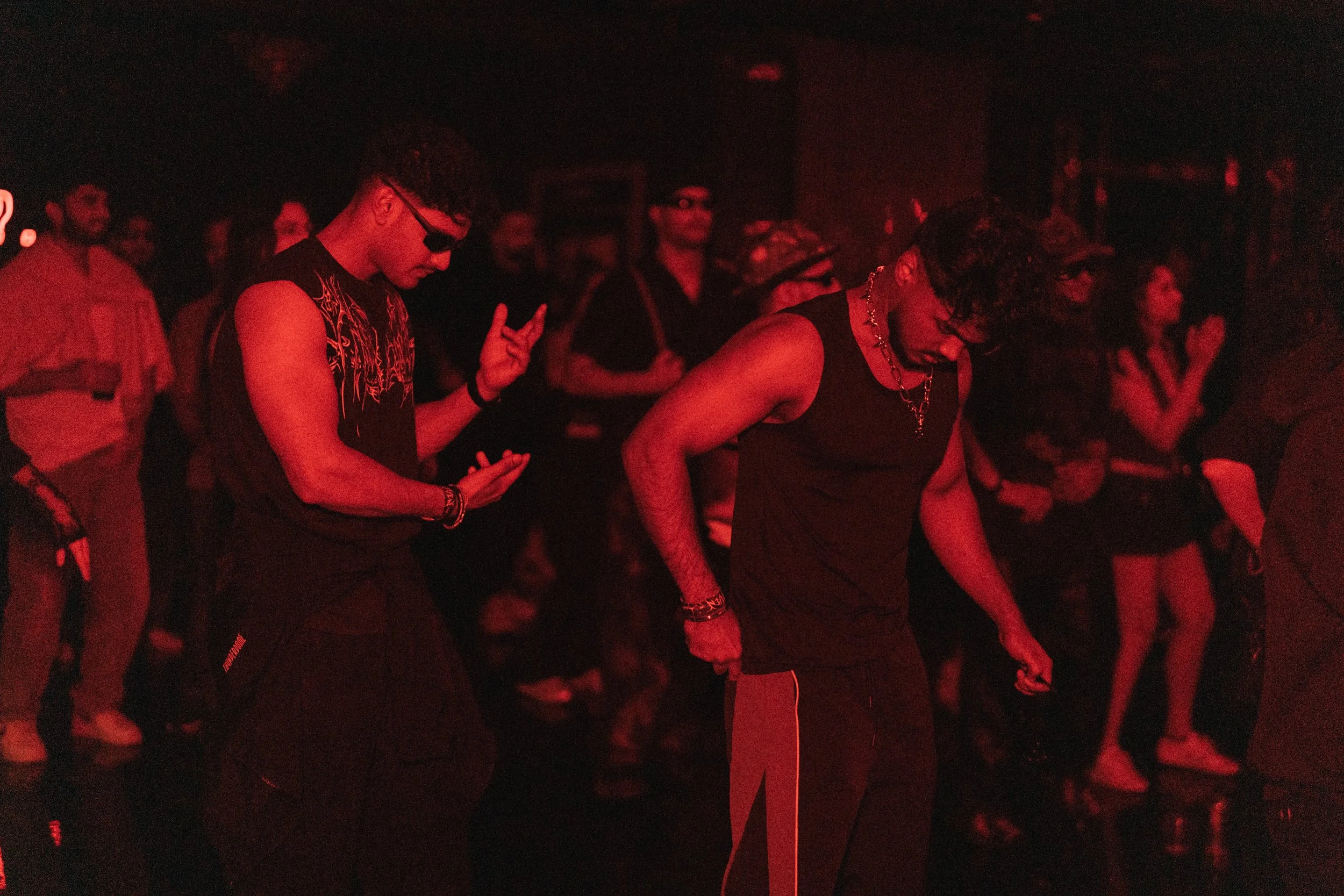 People dancing at a concert or club, illuminated by red lighting, with some wearing sunglasses and casual outfits.