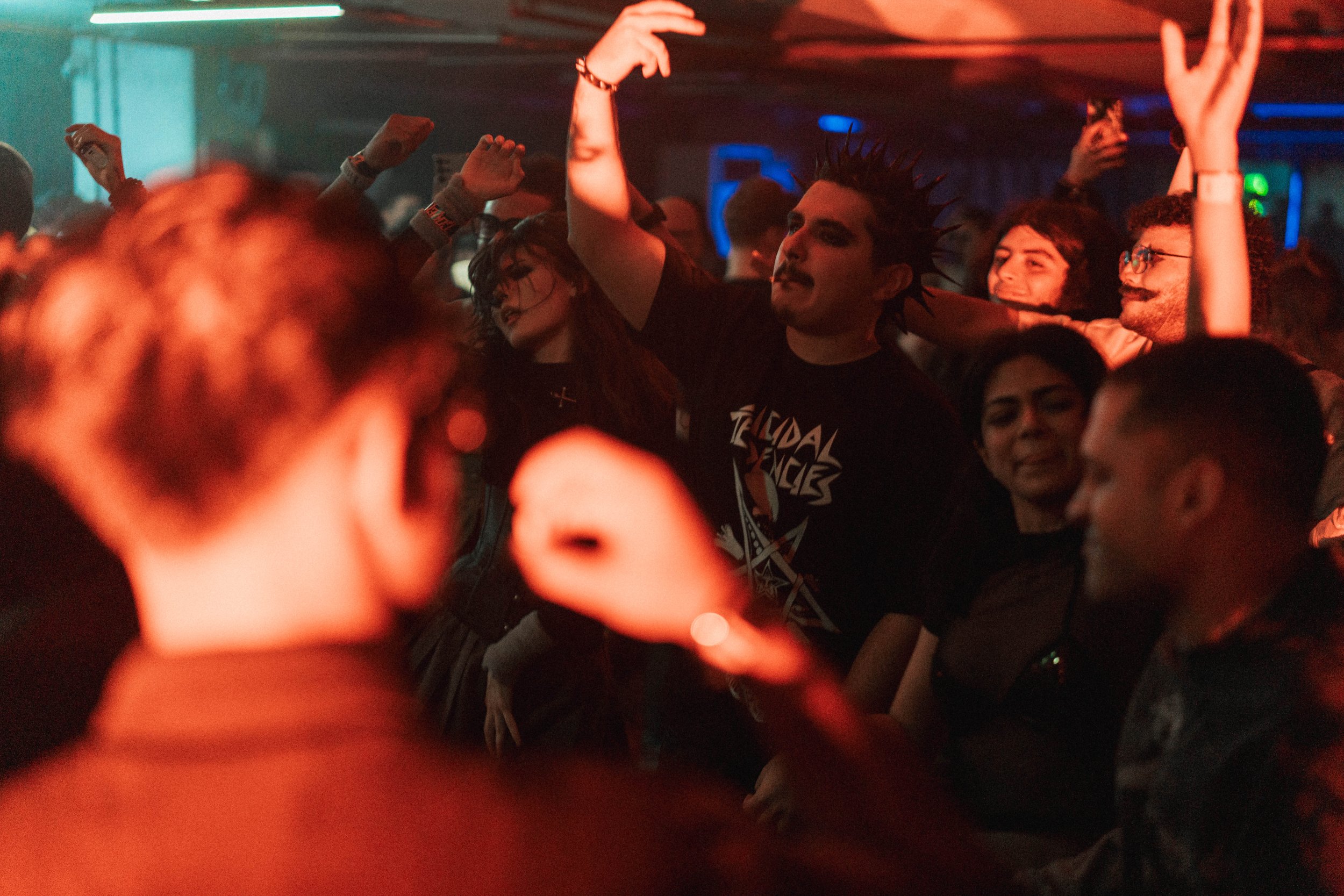 Crowd of young people enjoying music at a concert, some with hands raised, in a dimly lit indoor venue.