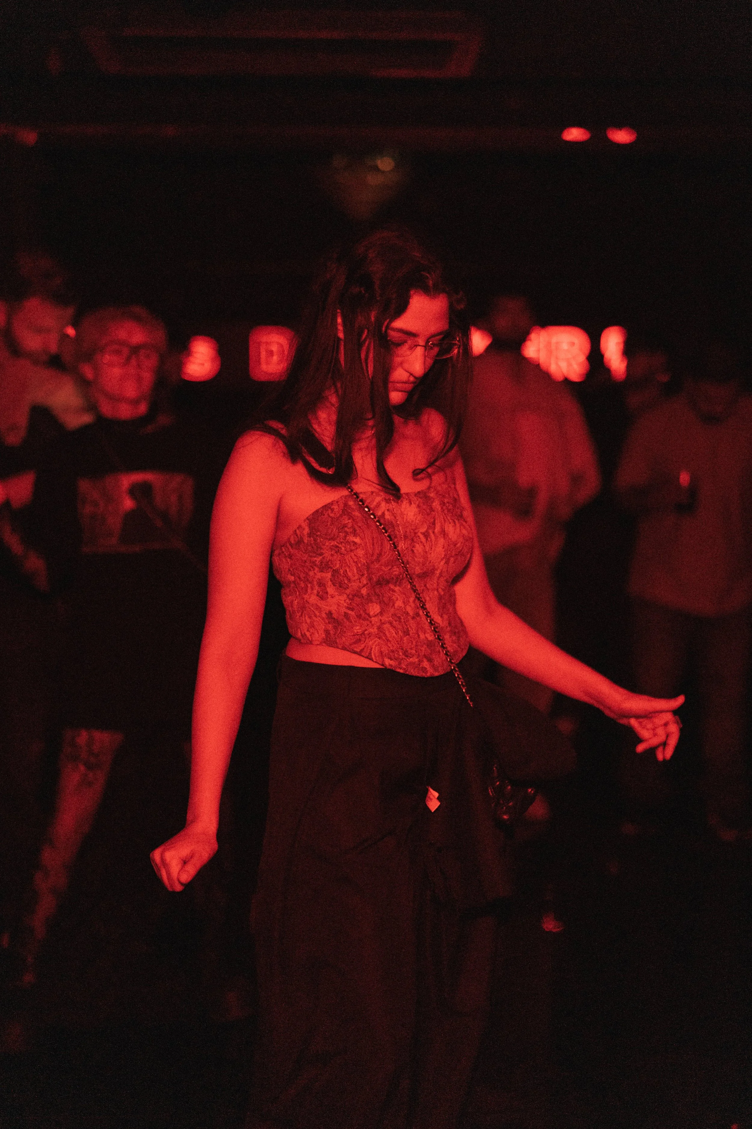A woman with dark hair wearing glasses, a patterned sleeveless top, and black pants dancing at a venue with red lighting, with several other people in the background.
