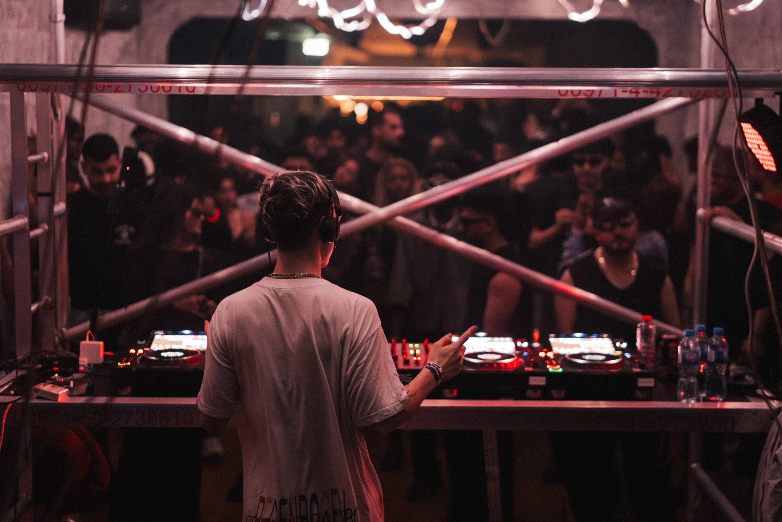 A DJ wearing a white shirt and headphones is performing at a nightclub, facing a crowd of people dancing and enjoying the music. The scene is dimly lit with red and pink lighting.