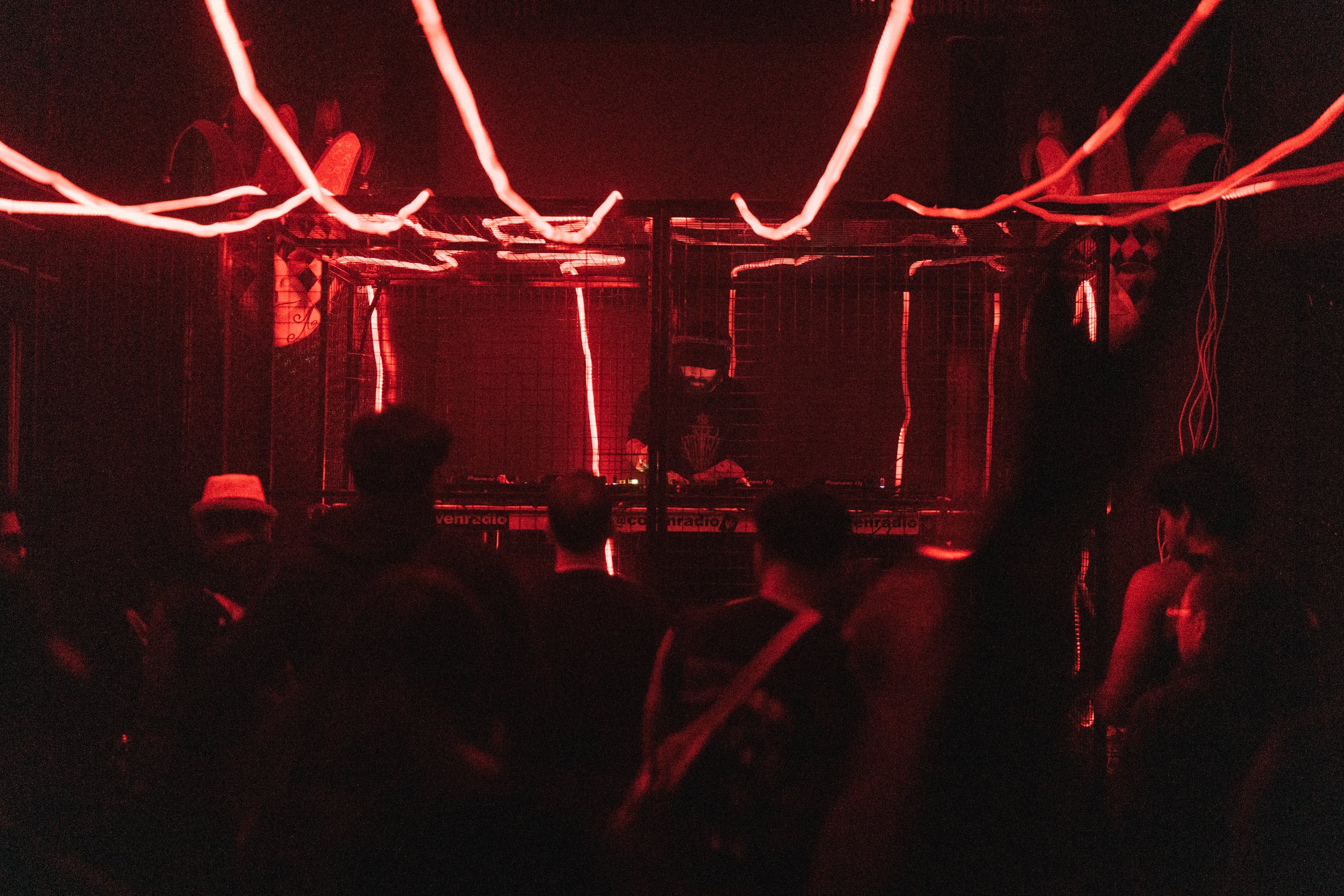 A DJ performs in a dark nightclub illuminated by red neon lights, with a group of people dancing and enjoying the music.
