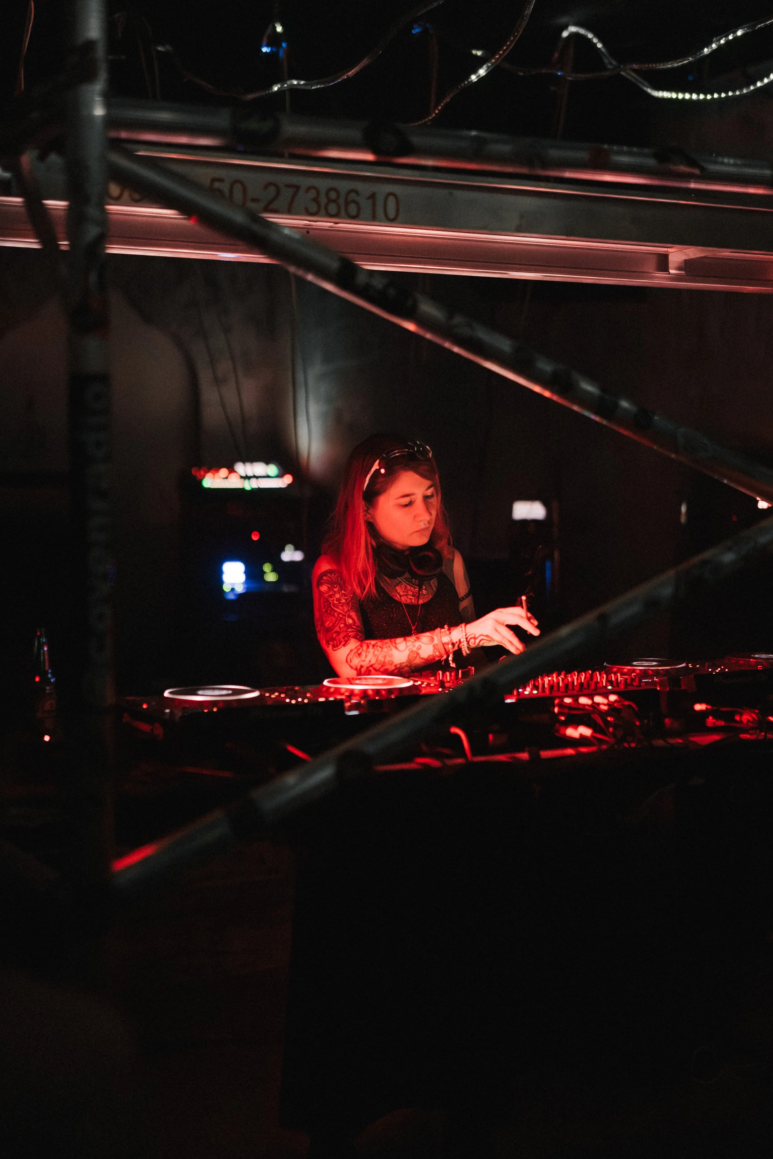A female DJ performing in a dimly lit club with red lighting, using DJ equipment behind a metal barrier.