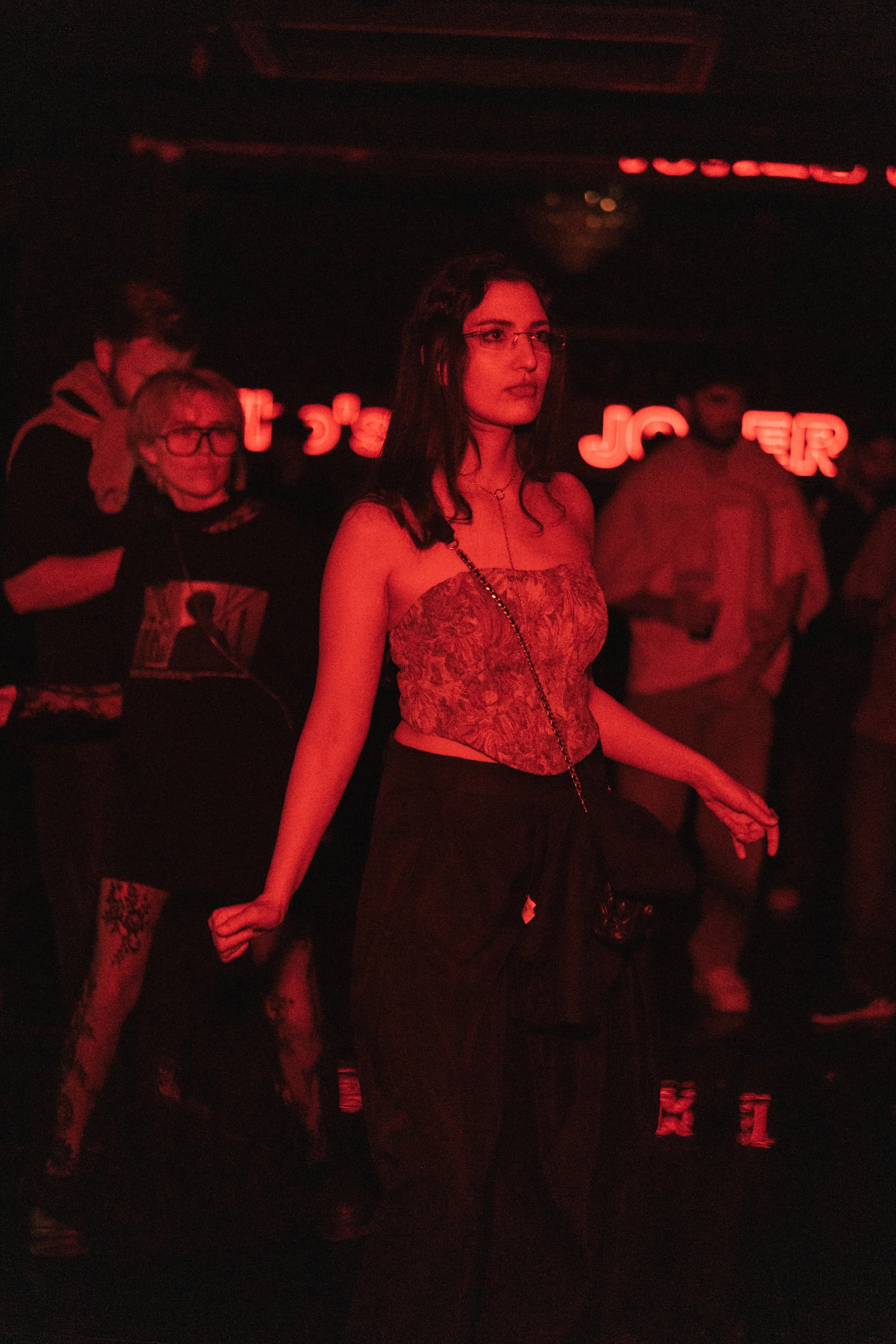 A woman in a strapless top and black pants dancing at a nightclub with red lighting, with several other people in the background and neon lights on the wall.