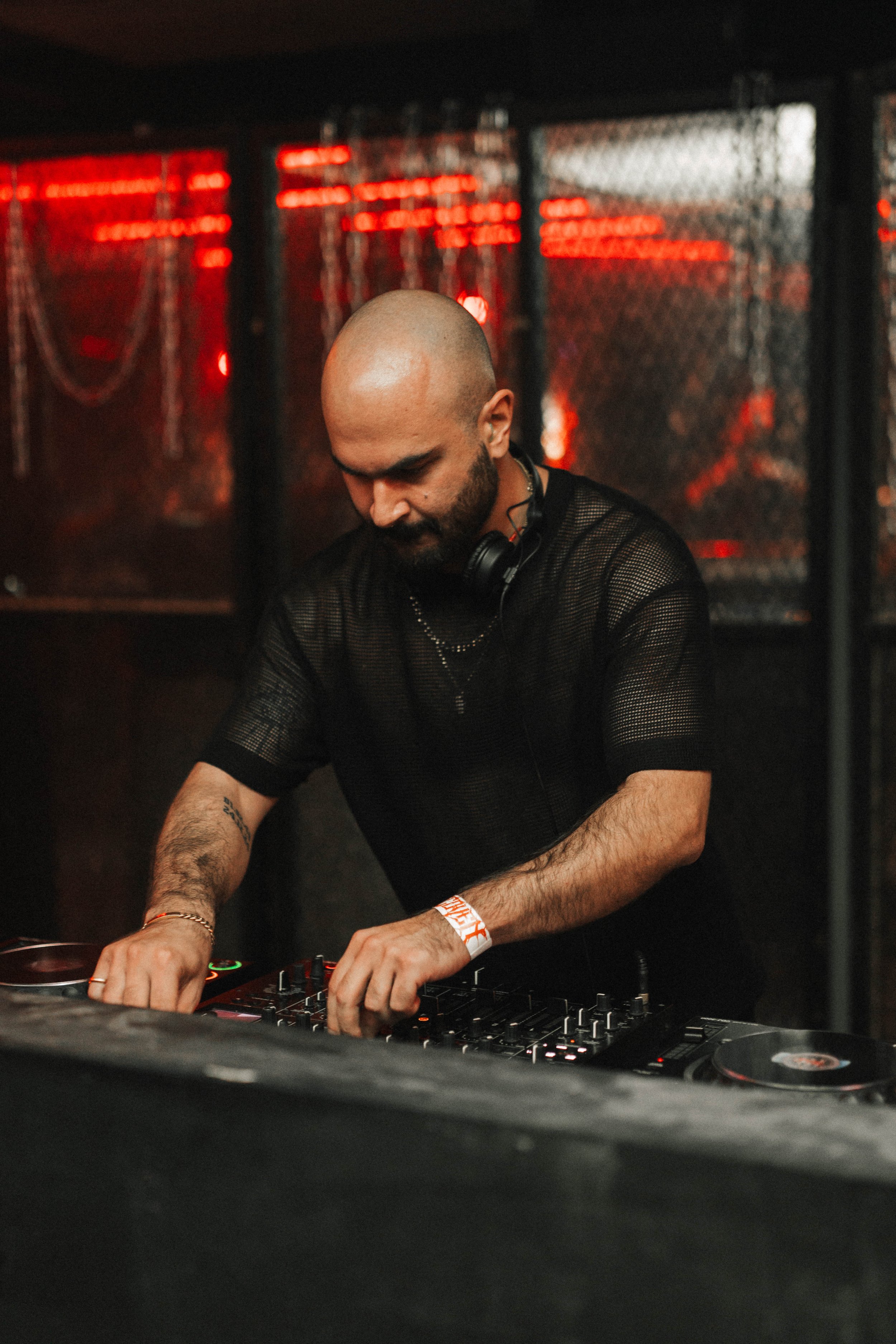 A male DJ with a shaved head and beard mixing music at a nightclub, wearing a black mesh shirt and headphones around his neck, with red lighting and metal fencing in the background.