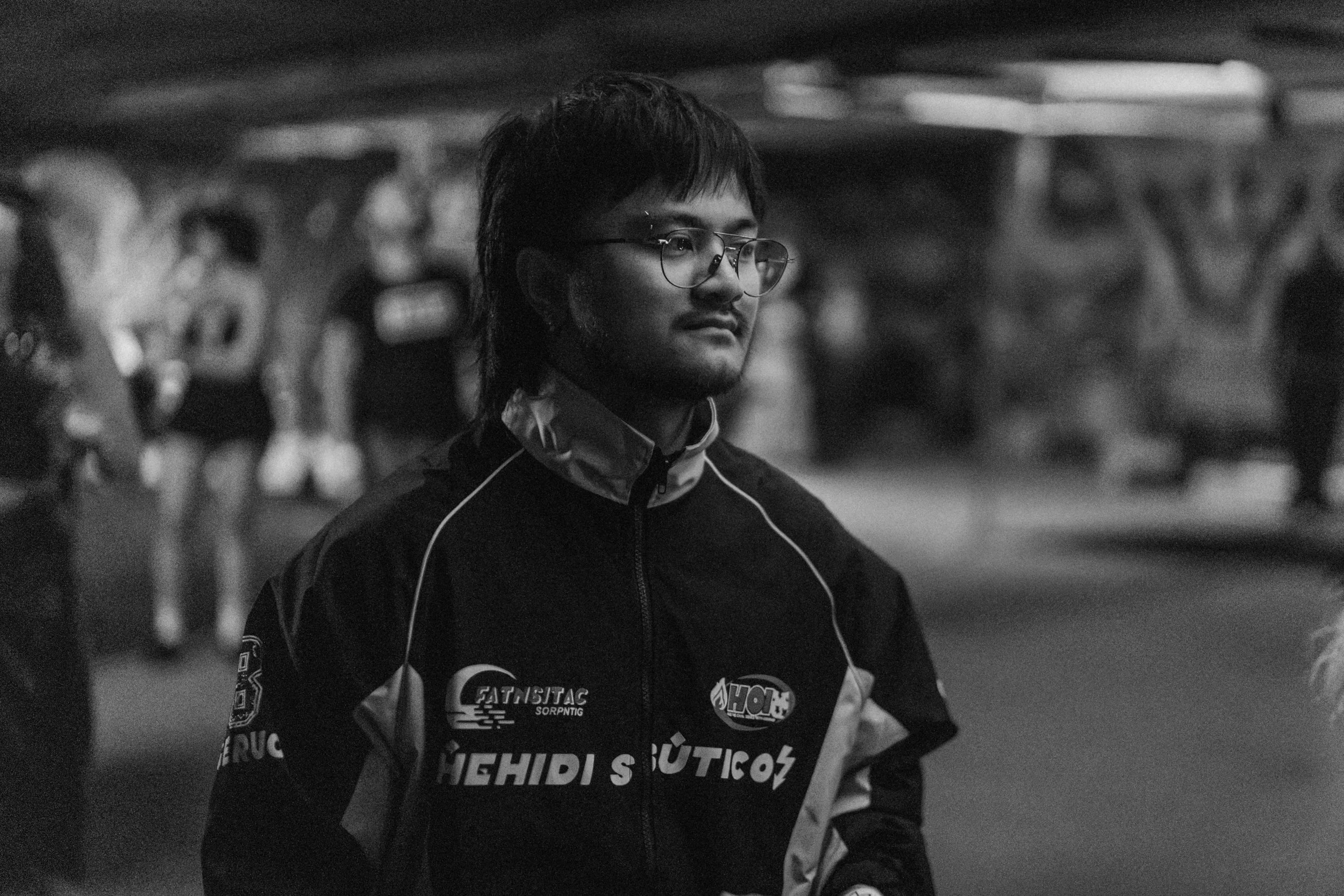Black and white photo of a man wearing glasses and a sports jacket, standing outdoors at night, with blurred people in the background.