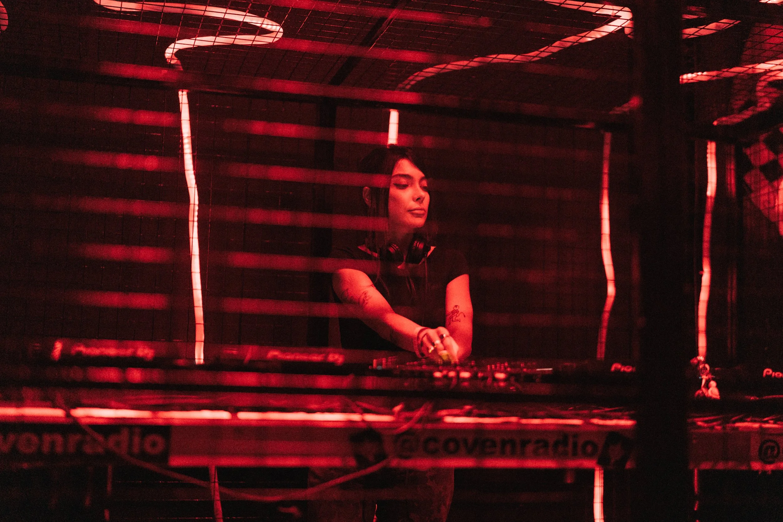 A female DJ performing behind a red-lit DJ booth, focused on her equipment in a dark environment.
