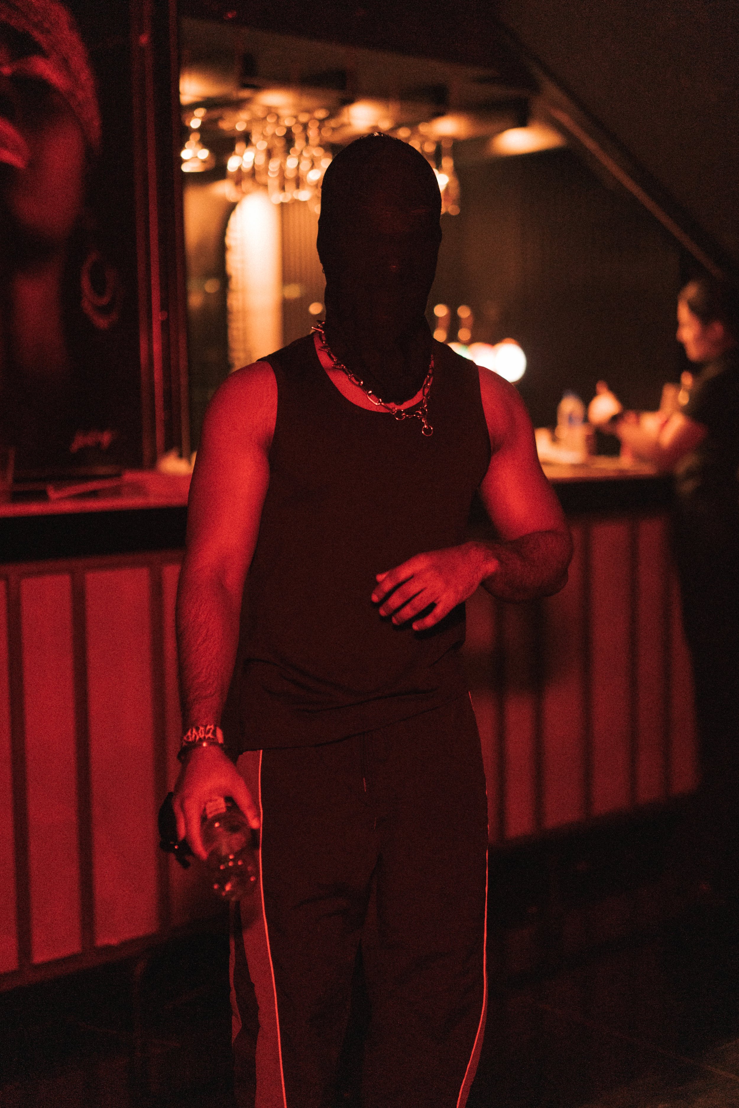 A person with dark skin is standing in a dimly lit bar or nightclub, wearing a sleeveless black top, pants with white stripes down the sides, and some jewelry including a chain necklace and bracelet. They are holding a bottle in one hand and appear t