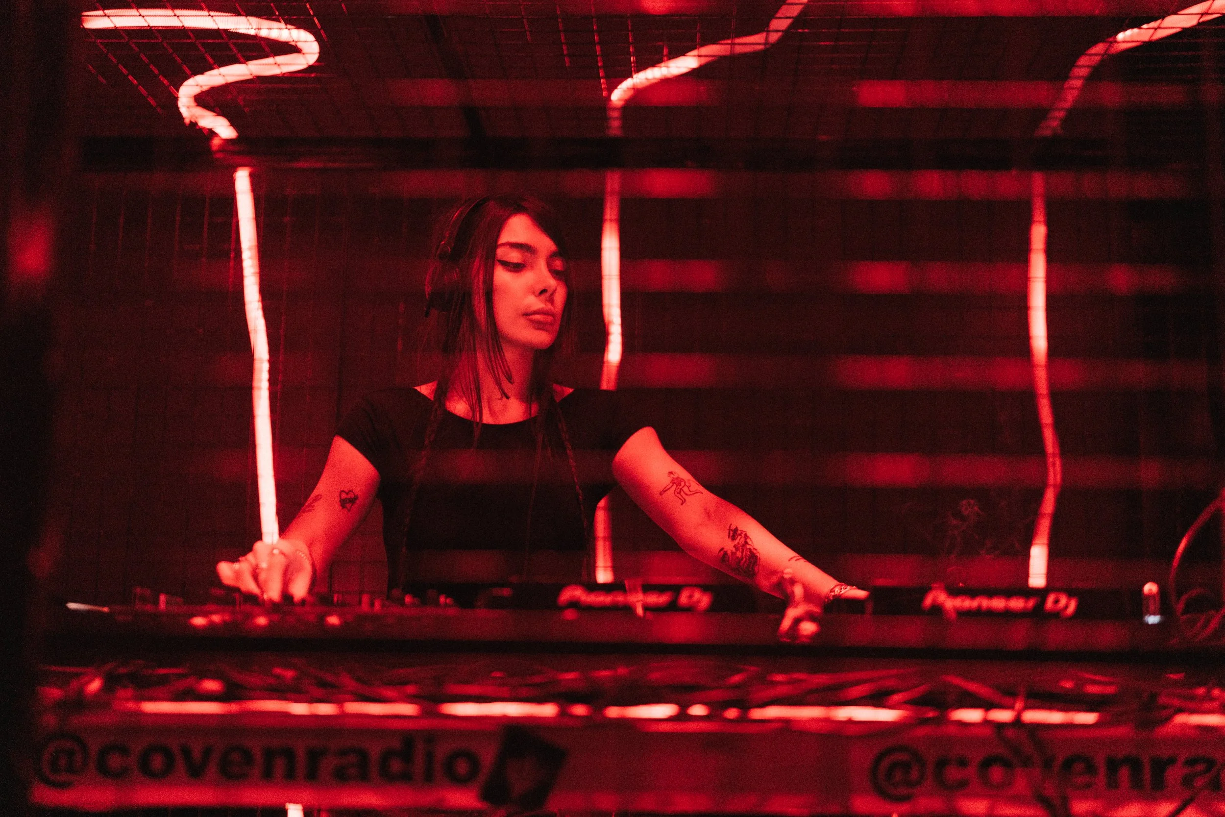 A female DJ with tattoos on her arms is performing at a club, illuminated by red neon lights.