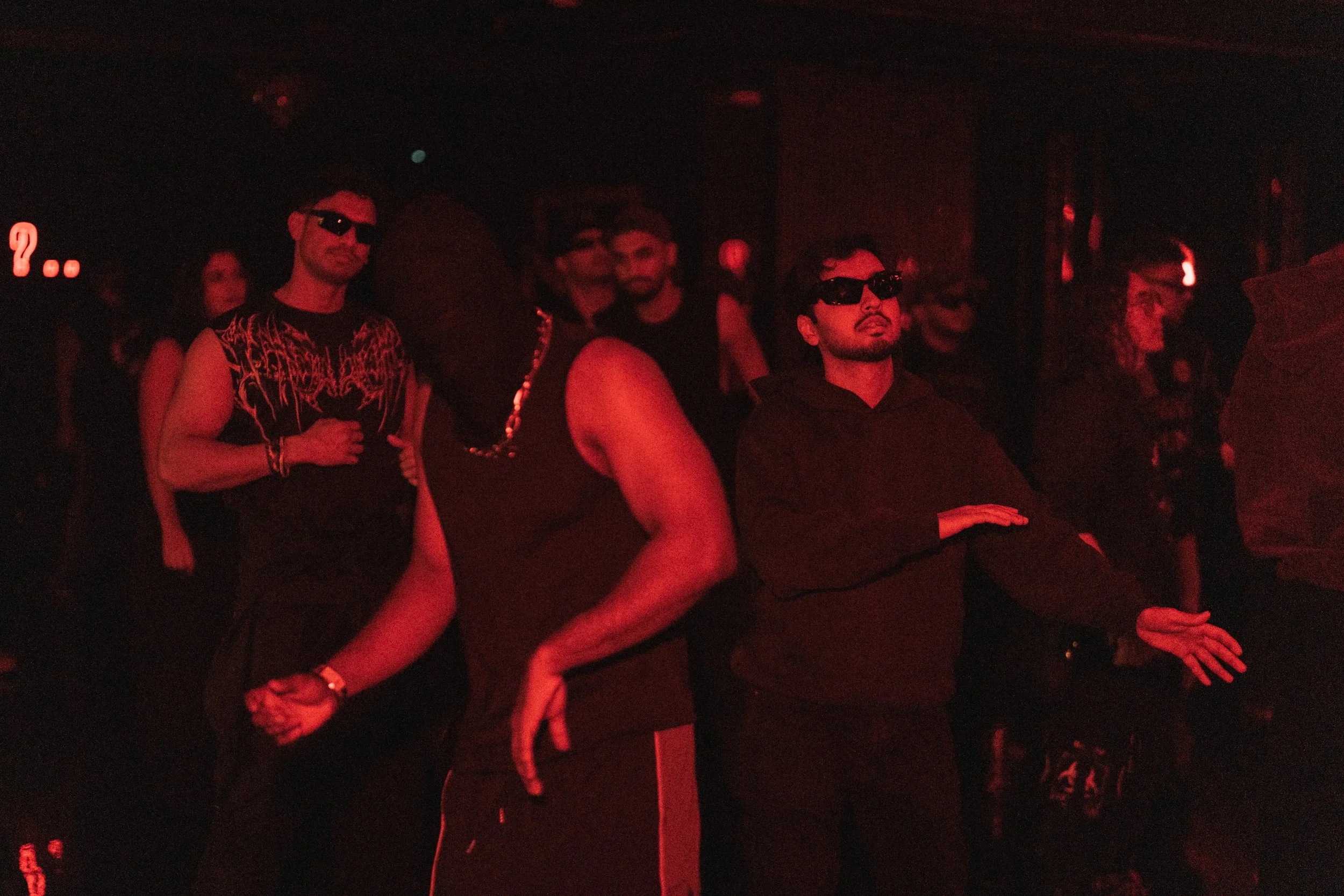 A group of people at a concert or nightclub, wearing dark clothing and sunglasses, enjoying the music in a dimly lit environment with red lighting.