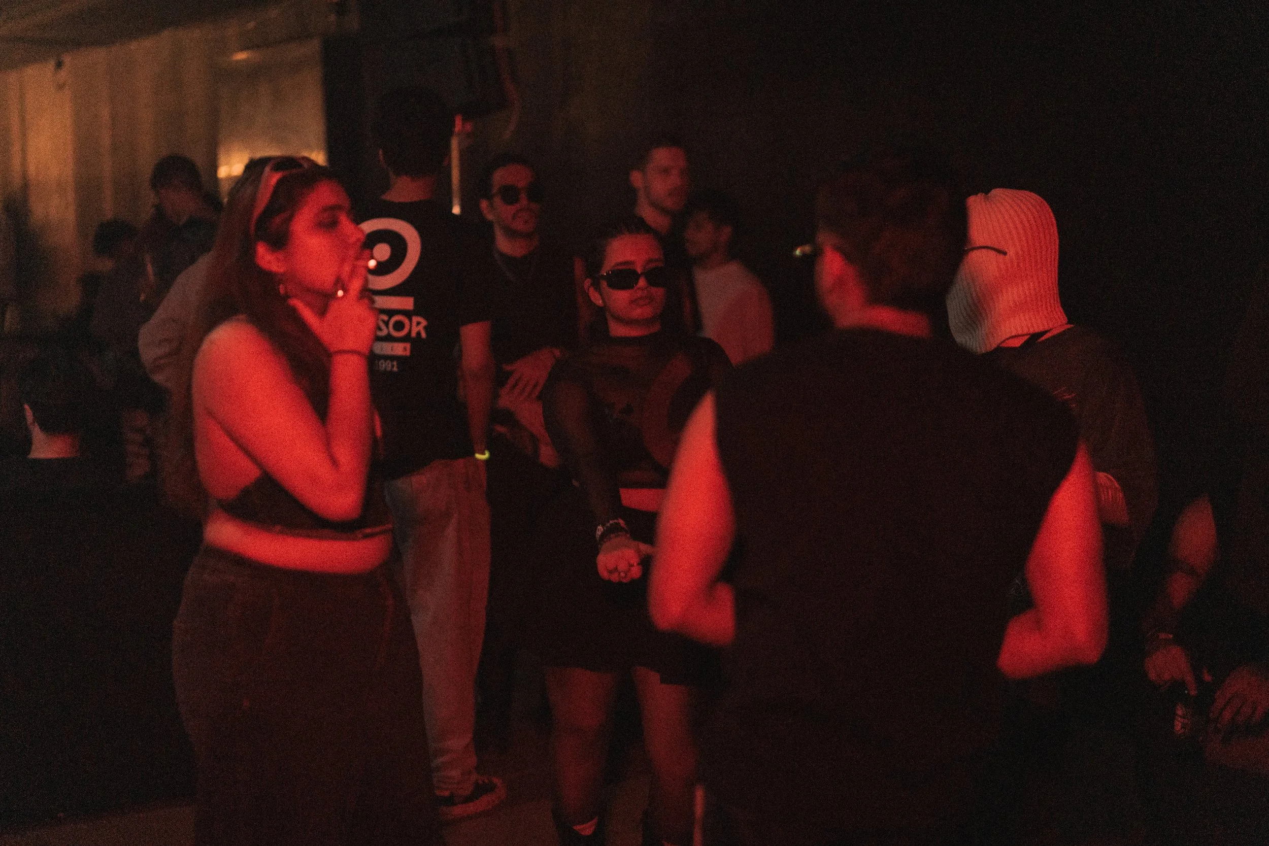 Group of young people at a dark, dimly lit party or club, some wearing dark clothing and sunglasses, engaged in conversation.