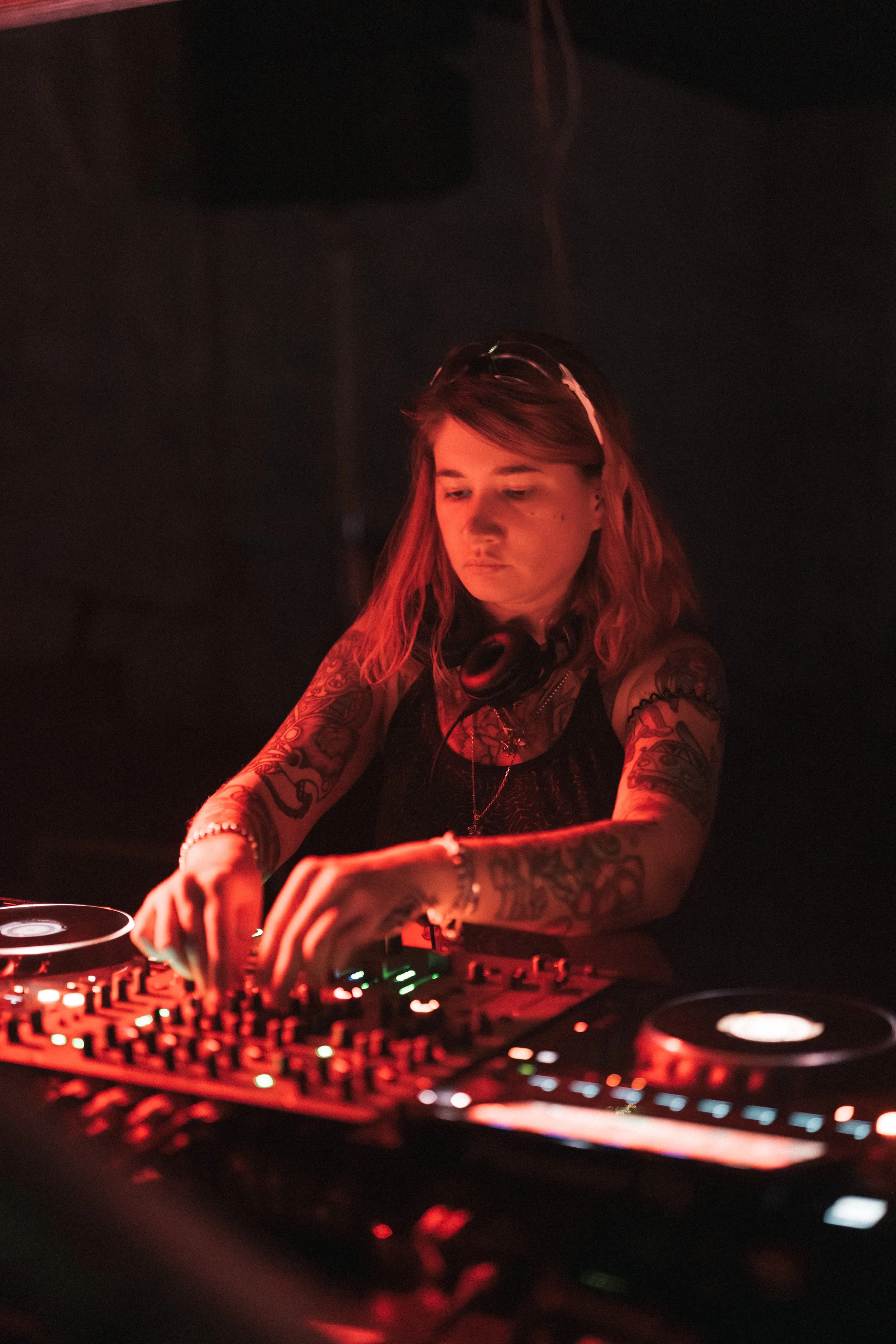 A female DJ with tattoos, wearing headphones around her neck, appears focused as she operates a DJ mixer and turntables in a dimly lit environment with red lighting.