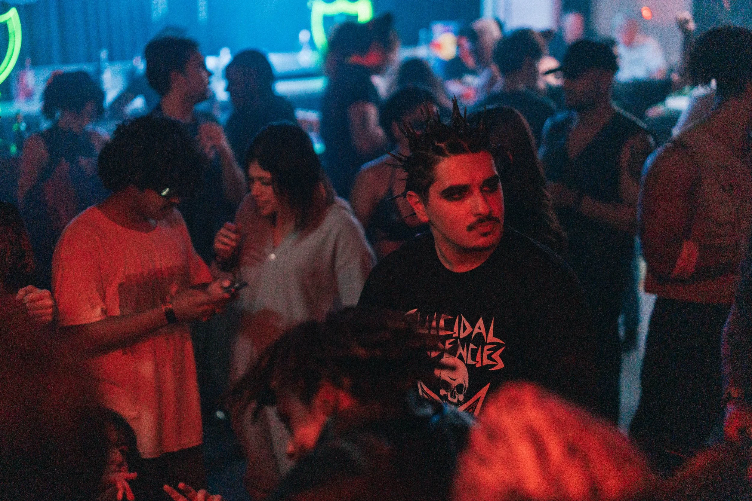 A person with black spiked hair and makeup resembling a punk or goth style, wearing a black T-shirt with a graphic and text, in a crowded, dimly-lit nightclub with blue and red lighting.