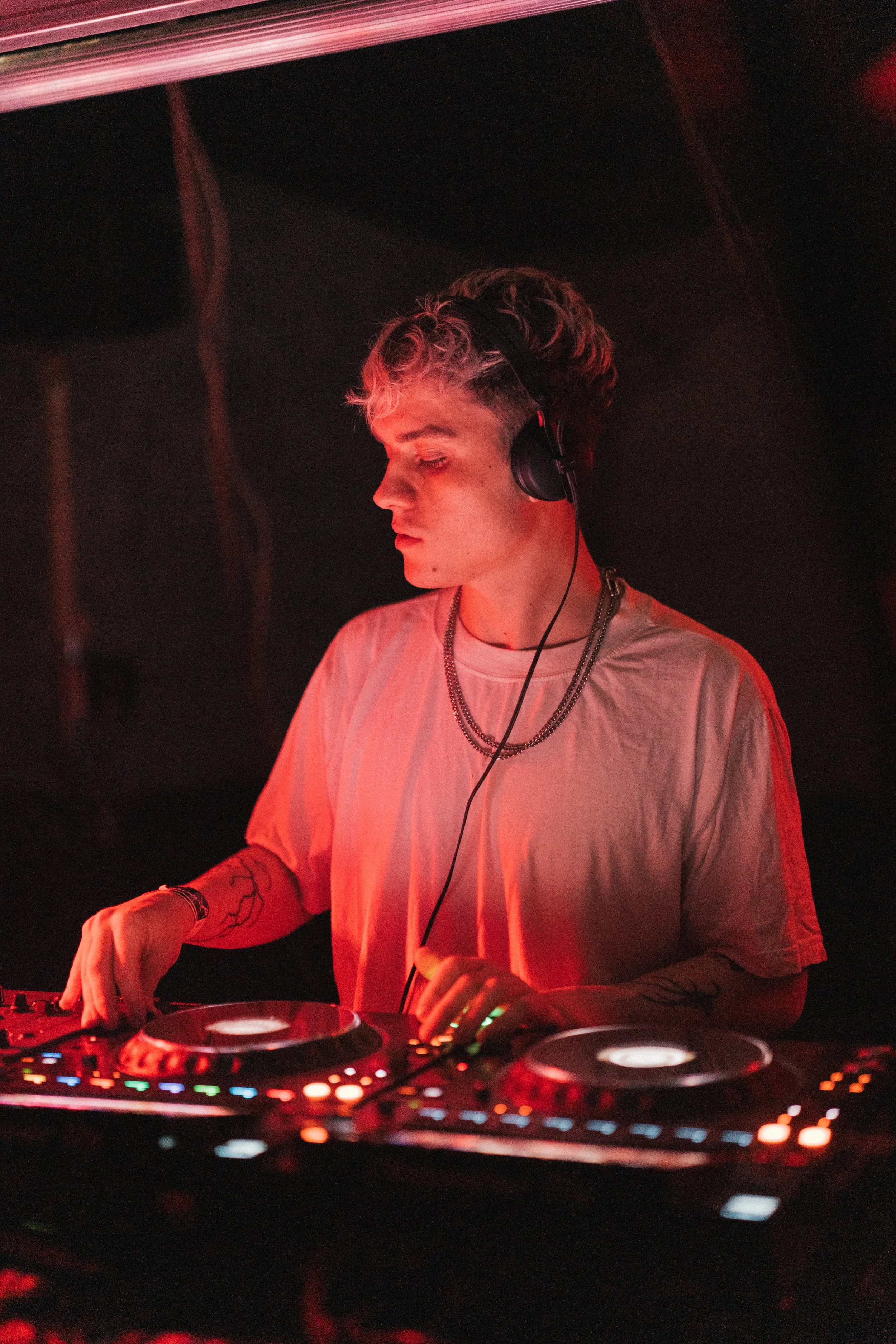 A young male DJ with blonde curly hair, wearing headphones, a white t-shirt, and layered necklaces, is mixing music at a DJ console in a dimly lit setting with red lighting.