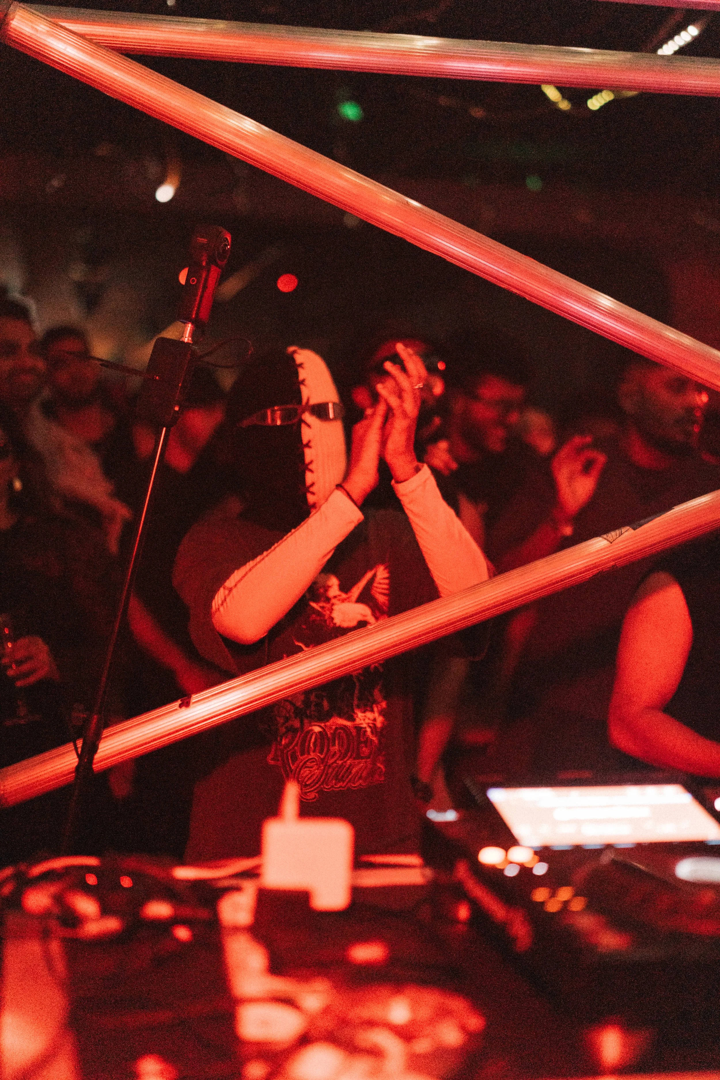 Person wearing a mask and headphones, standing behind DJ equipment at a dimly lit, red-colored concert or club event, with a crowd of people in the background.