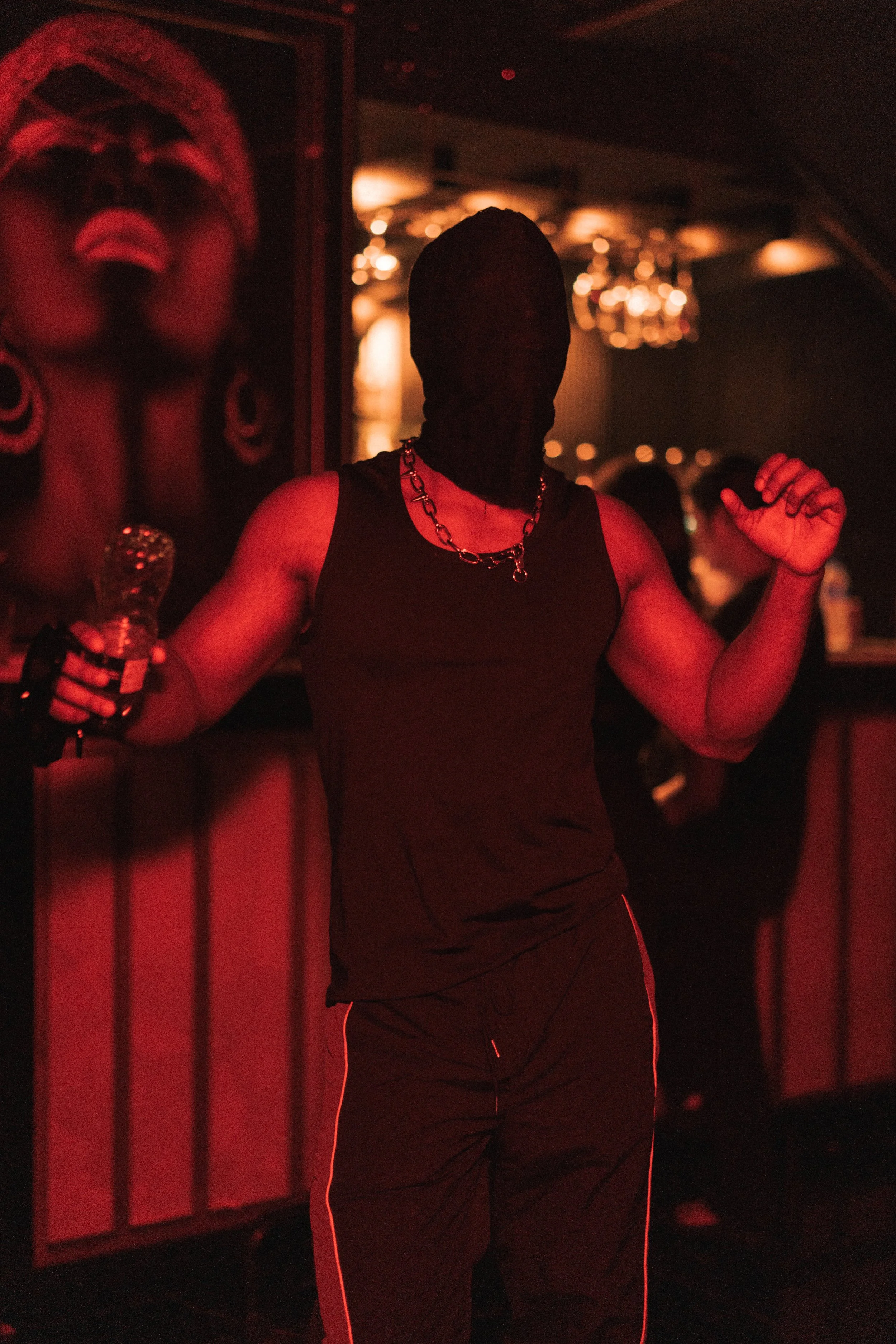 A person wearing a black sleeveless shirt, a gold chain necklace, and black pants standing in a dimly lit bar or club with red lighting. The person's face is obscured by a black mask or hood, and they are holding a bottle of beer in one hand.