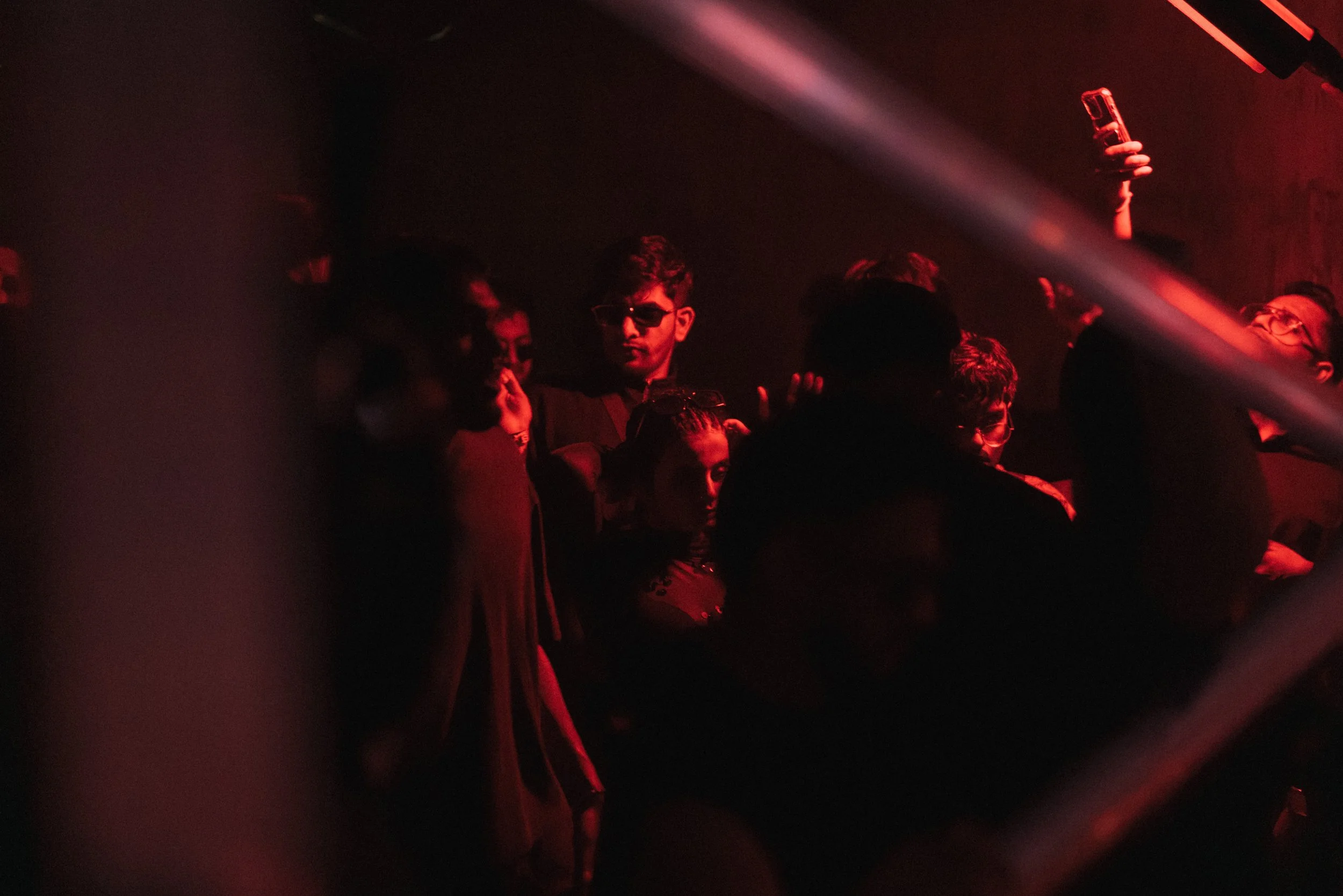 Group of people at a dark nightclub with red lighting, some wearing sunglasses, some taking photos or videos.