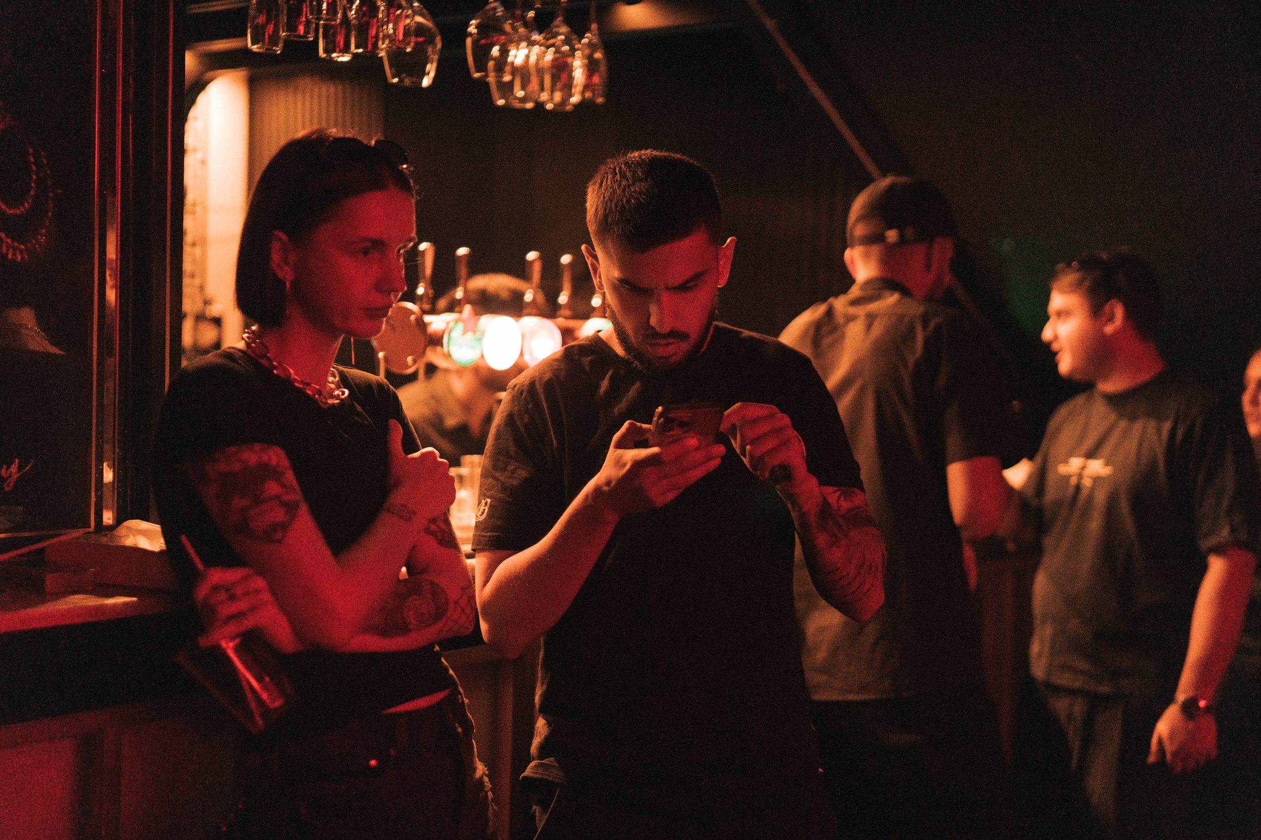People standing at a bar in a dimly lit nightclub, with some looking at their phones and drinking.