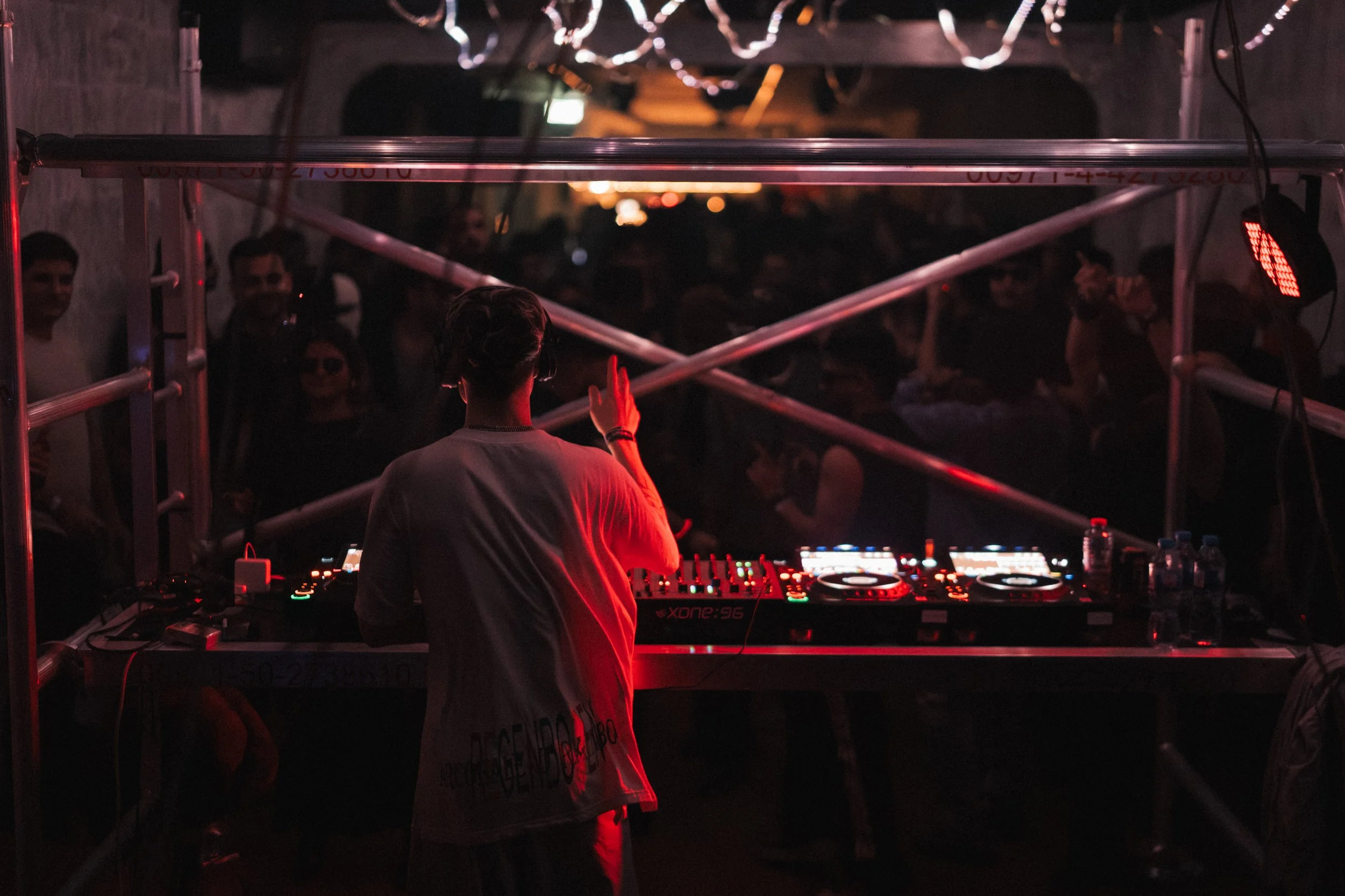 DJ performing at a nightclub with a crowd in the background, illuminated by red and purple lighting.