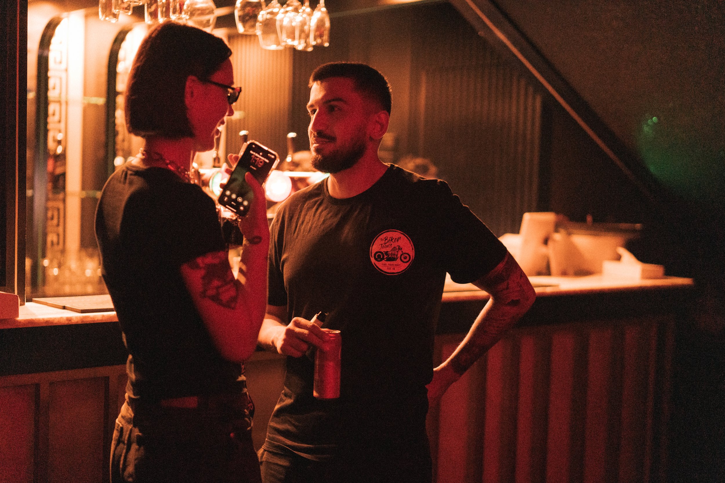 Two people talking at a bar with glasses hanging above them, in a dimly lit setting.
