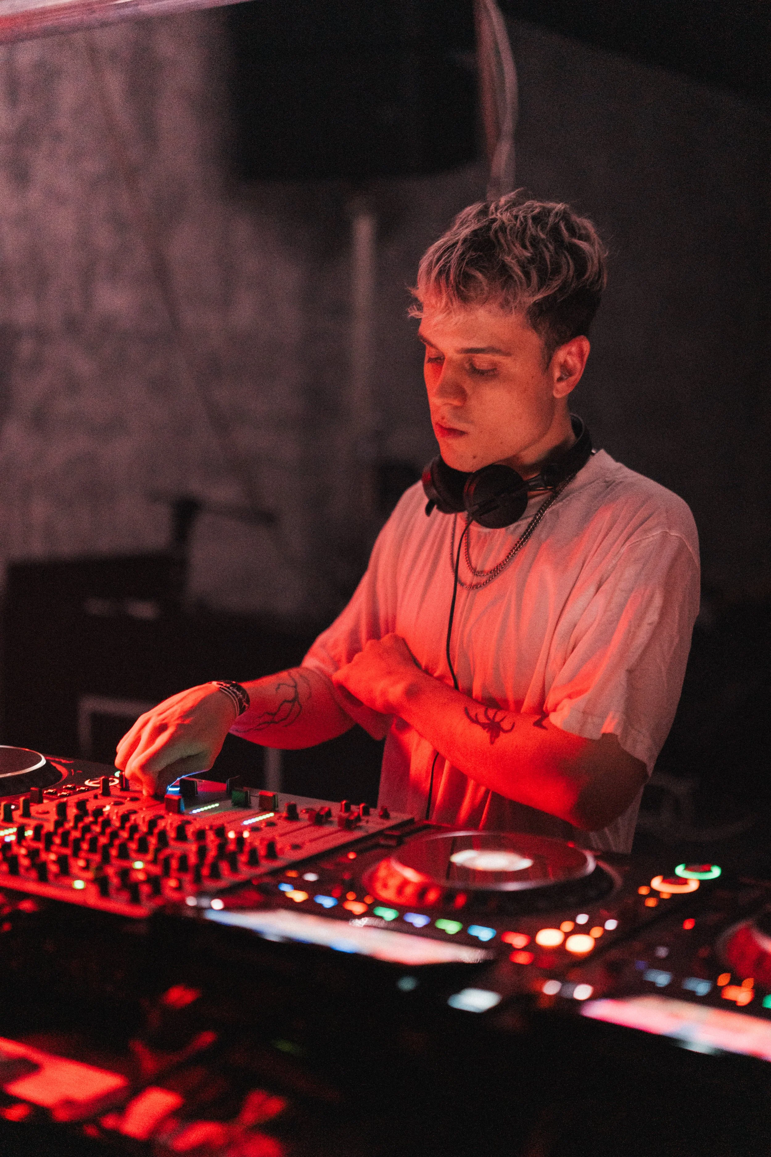 A young male DJ with tattoos on his arms, wearing a white t-shirt and headphones around his neck, is operating a professional DJ mixer at a party or club, with red lighting creating a vibrant atmosphere.