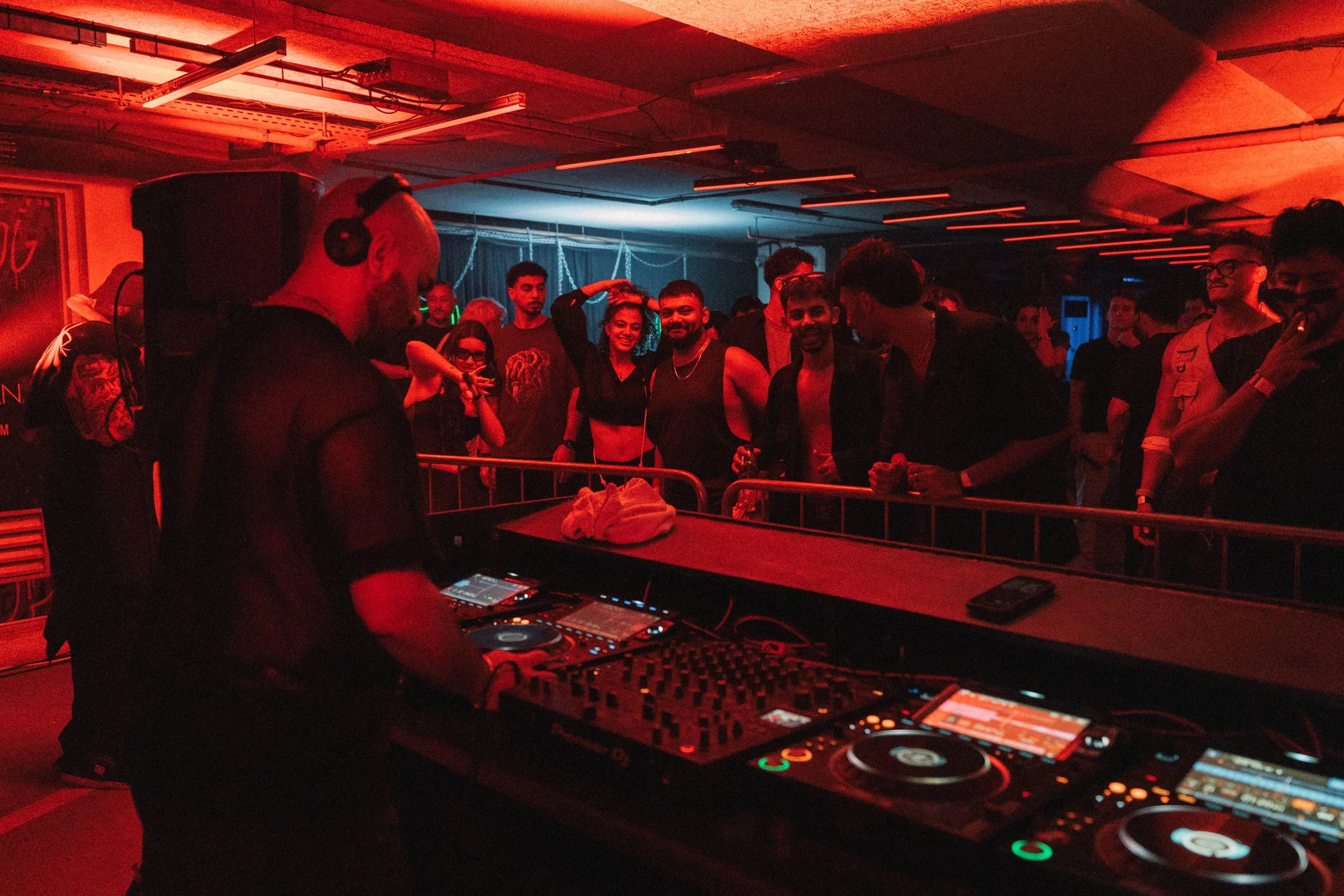A DJ with headphones on performing at a nightclub with a crowd of young adults dancing and enjoying the music, illuminated by red lighting.