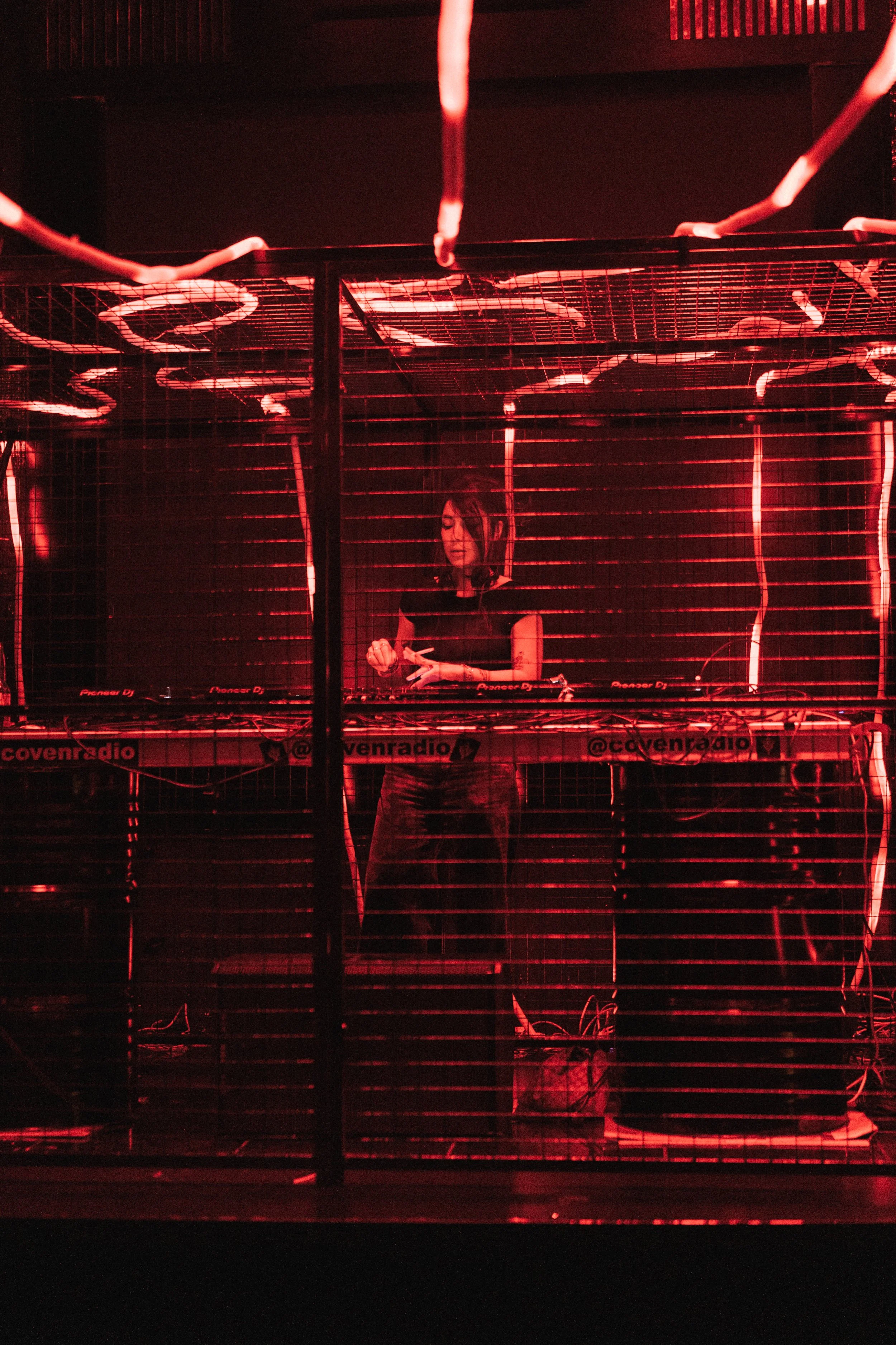 A female DJ performing behind a wire cage, illuminated by red neon lights at a club or concert.