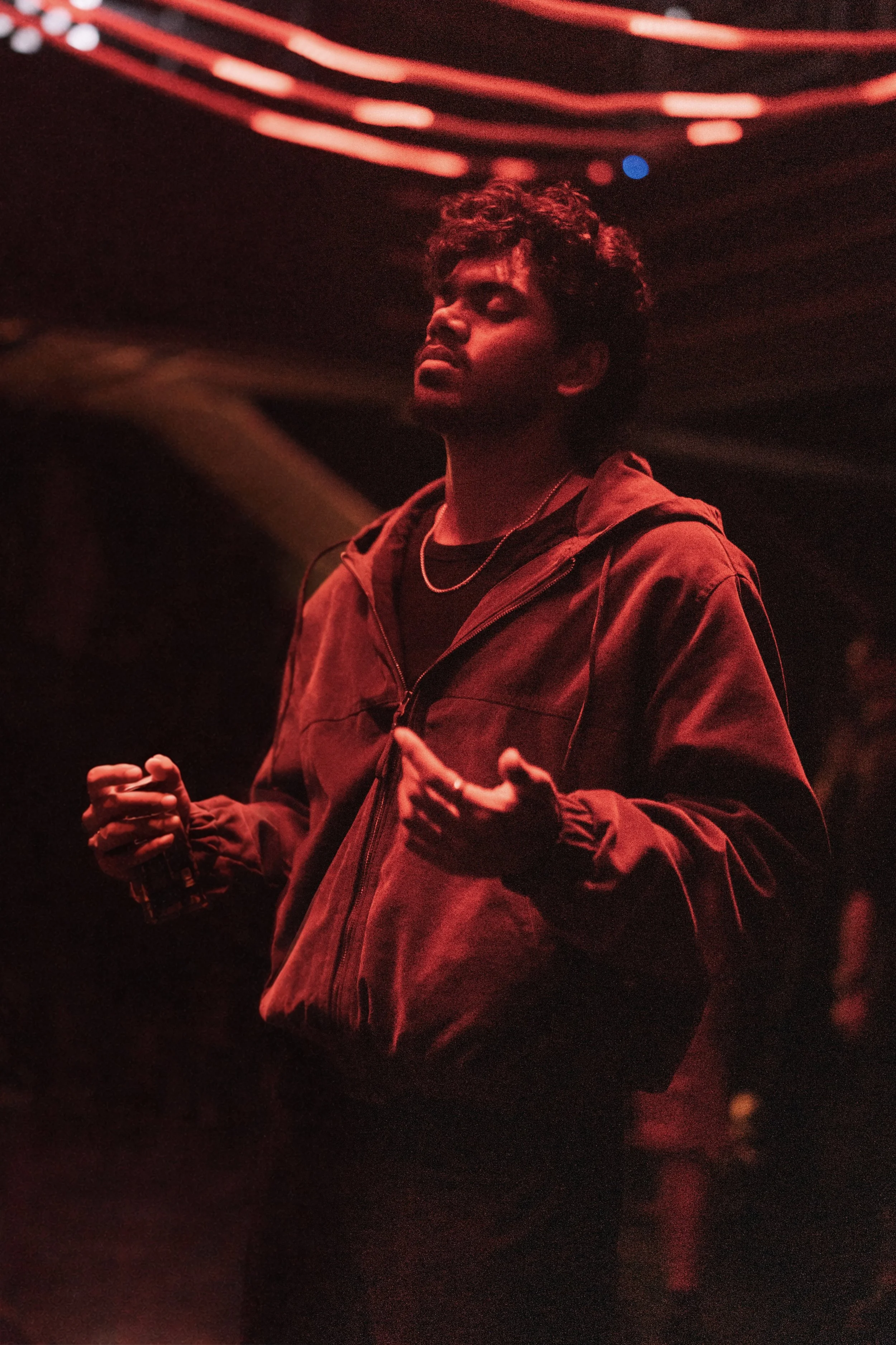 A young man with curly hair and a beard wearing a dark jacket and a silver necklace, standing in dim red lighting, holding a drink in one hand, appearing to be deep in thought or listening.
