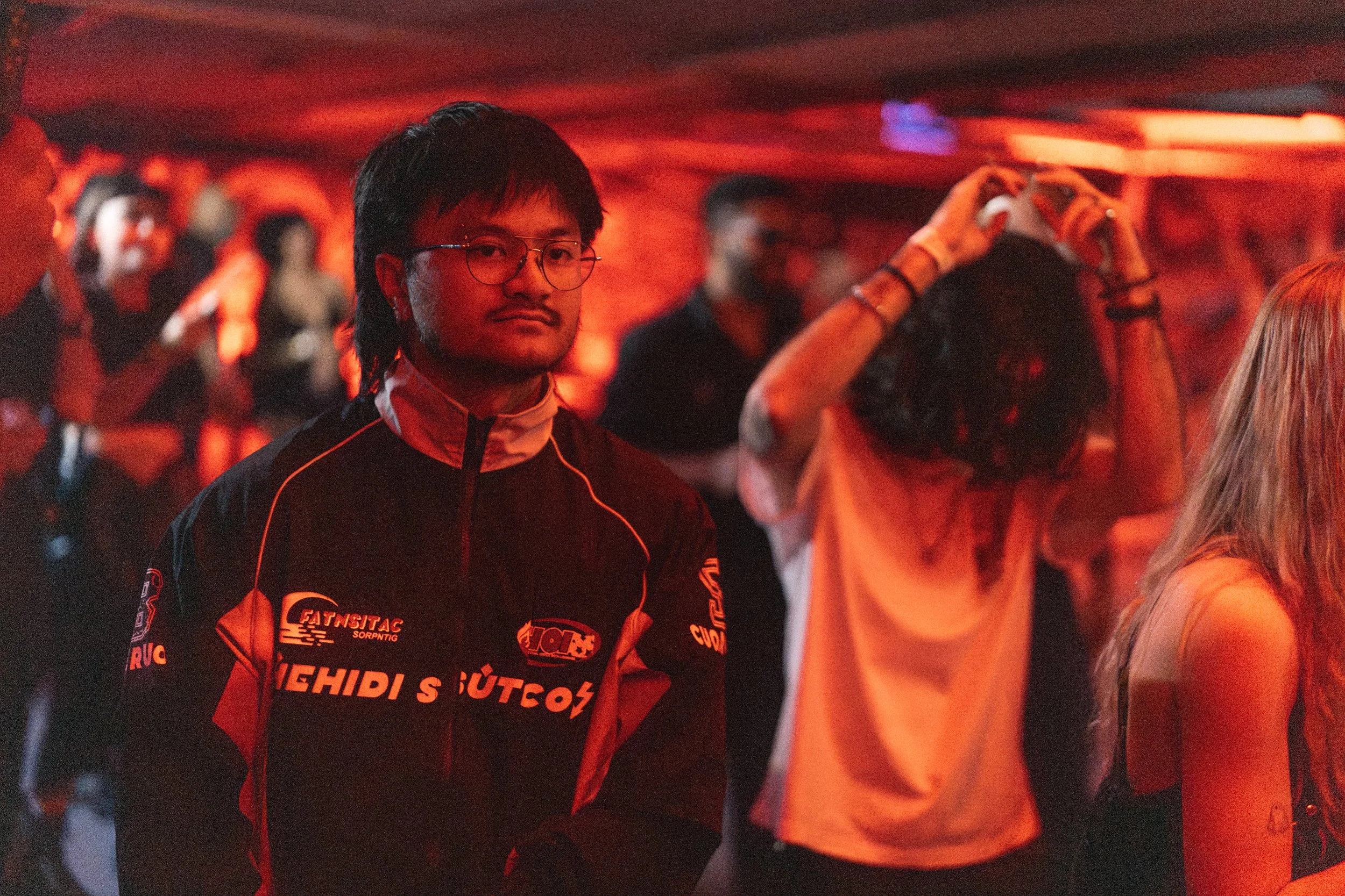 A young man wearing glasses and a racing jacket stands in a dimly lit room with red lighting, surrounded by other people, some of whom are dancing or taking photos.