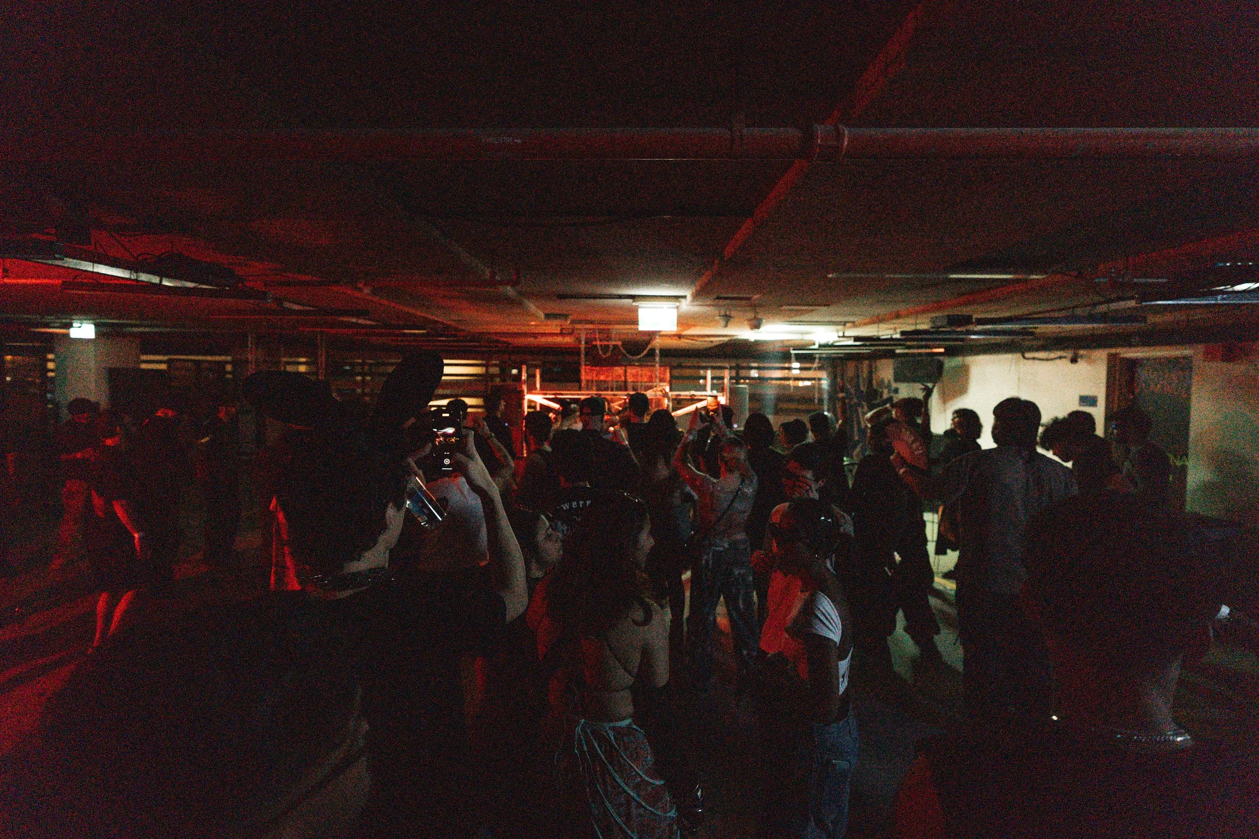 A crowd of people in a dark indoor space with red lighting, music equipment, and industrial piping on the ceiling, suggesting a concert or nightclub setting.