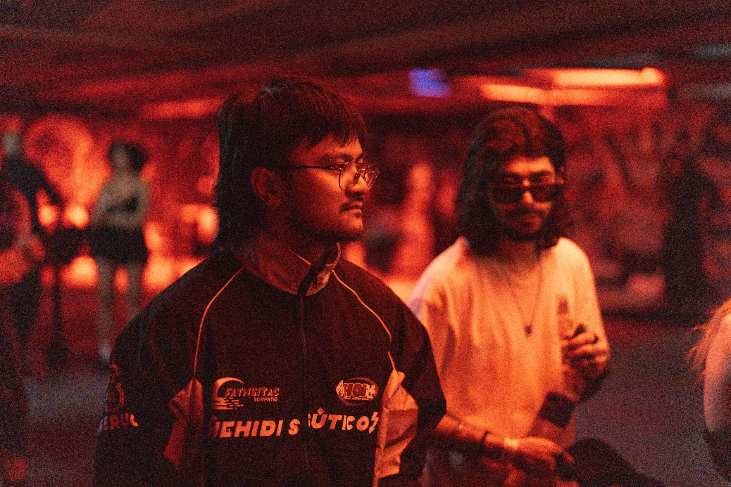 Two young men with dark hair and glasses standing in a dimly lit room with red lighting. The man on the left is wearing a sports jacket with various logos, and the man on the right is wearing a light-colored T-shirt, sunglasses, and a necklace.