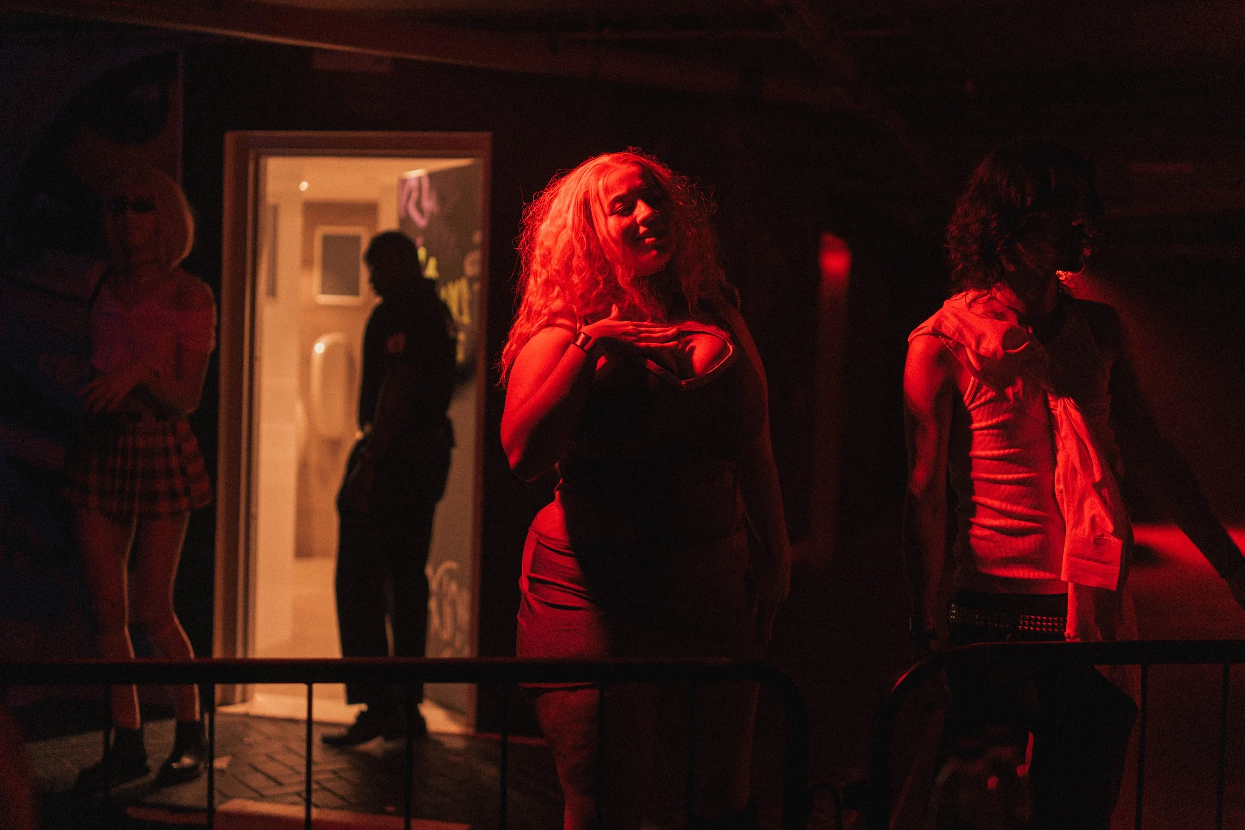 A group of people at a dimly lit, red-toned indoor event or club, with one woman in the center, gesturing or singing, and others standing around or near the doorway.