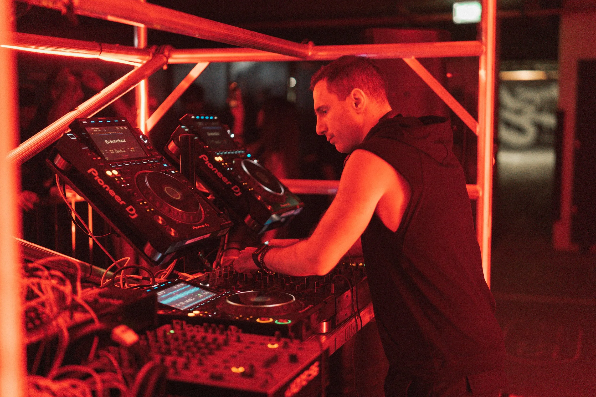 A DJ wearing a sleeveless black hoodie sets up equipment at a nightclub, illuminated by red lighting.