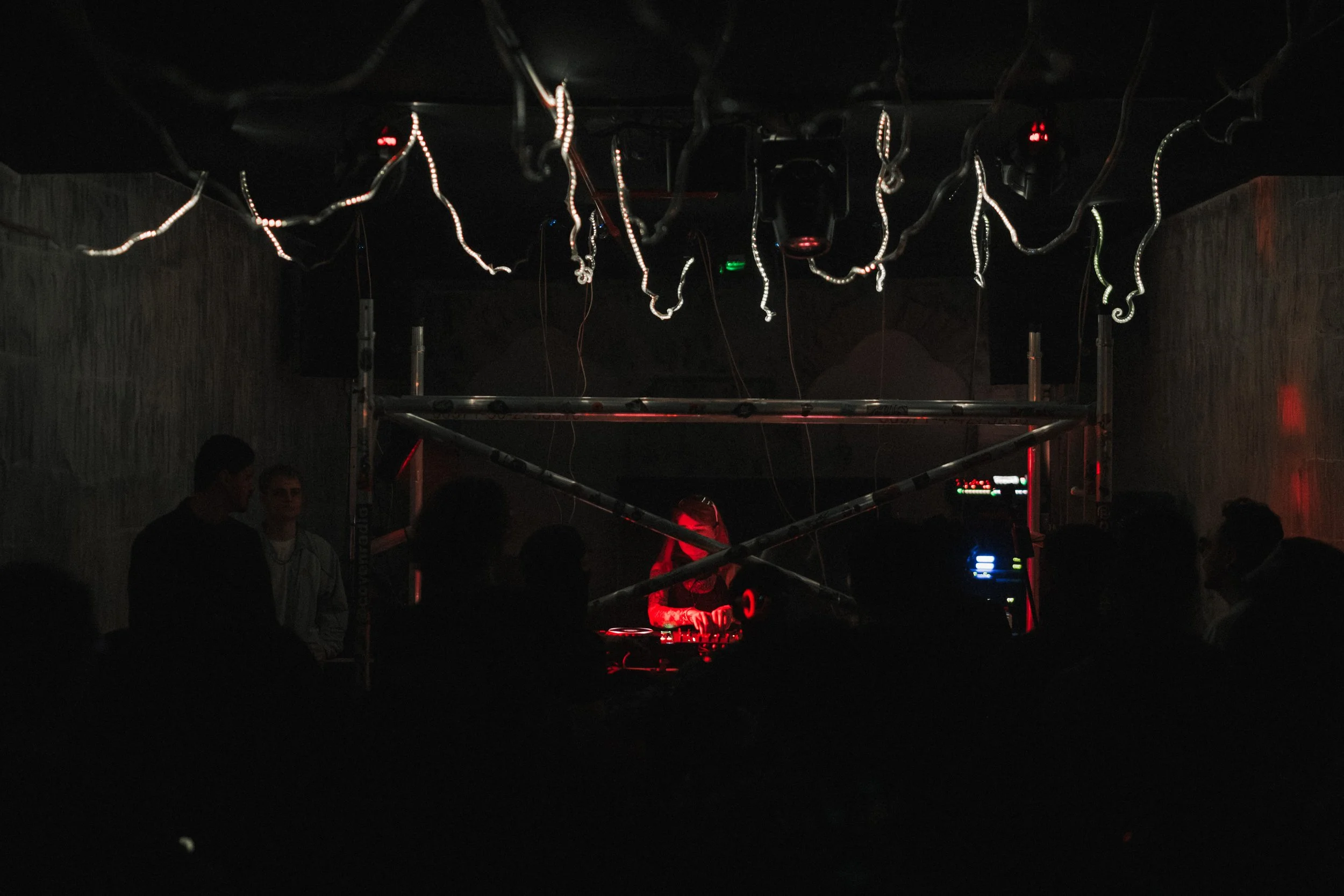 A DJ performing in a dark nightclub with people dancing and socializing. The DJ is illuminated by red and blue lights, and there are decorative light strips hanging from the ceiling.