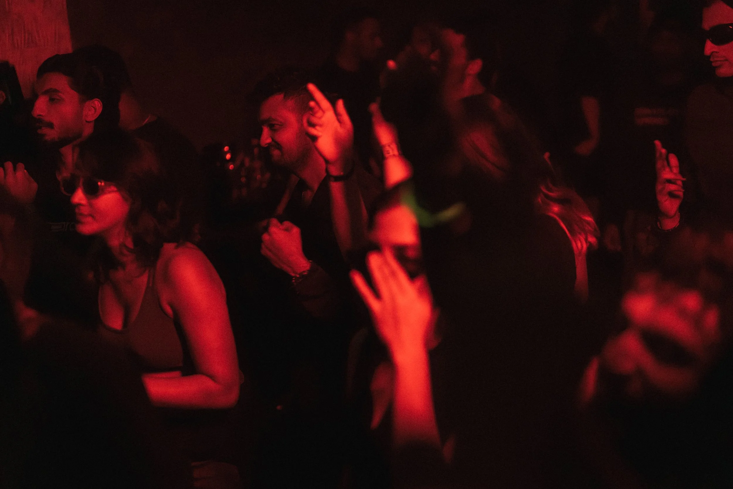 People dancing and enjoying music in a dark, red-lit nightclub or concert venue.
