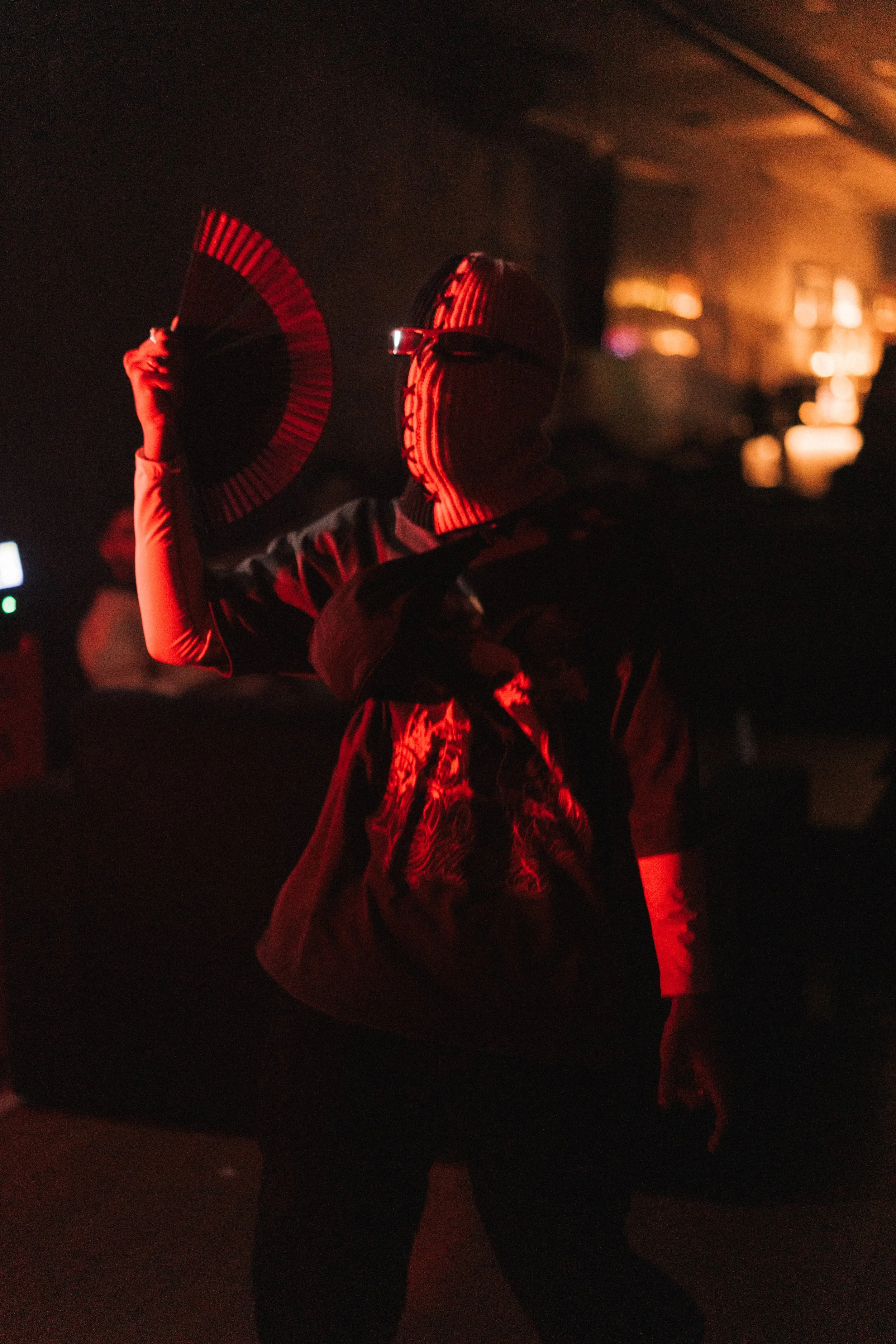 Person wearing a mask with horizontal stripes, holding a fan, in a dimly lit environment with red lighting, carrying a bag with an intricate pattern.