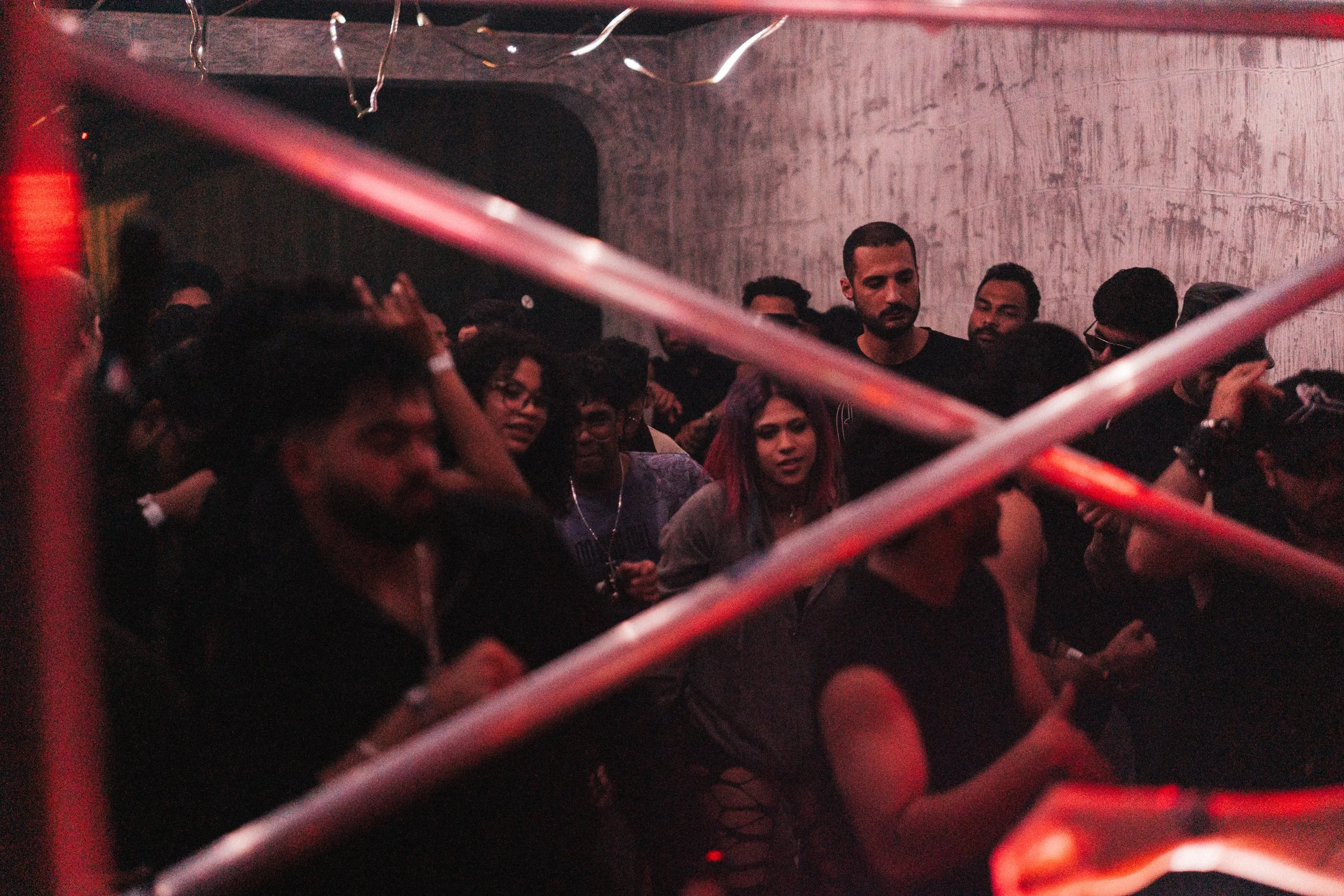 Crowd of people at a party or concert in a dimly lit room with red lighting, seen through metal bars.