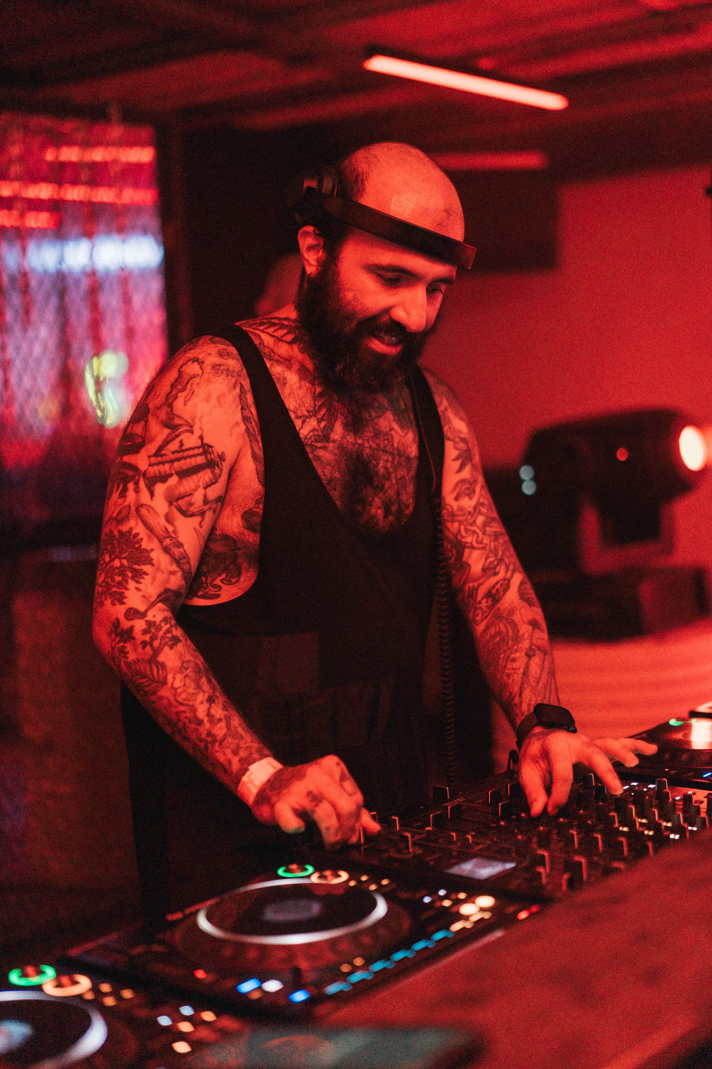 A DJ with tattoos on his arms, wearing a black tank top and headphones, playing music on DJ equipment in a red-lit club or venue.