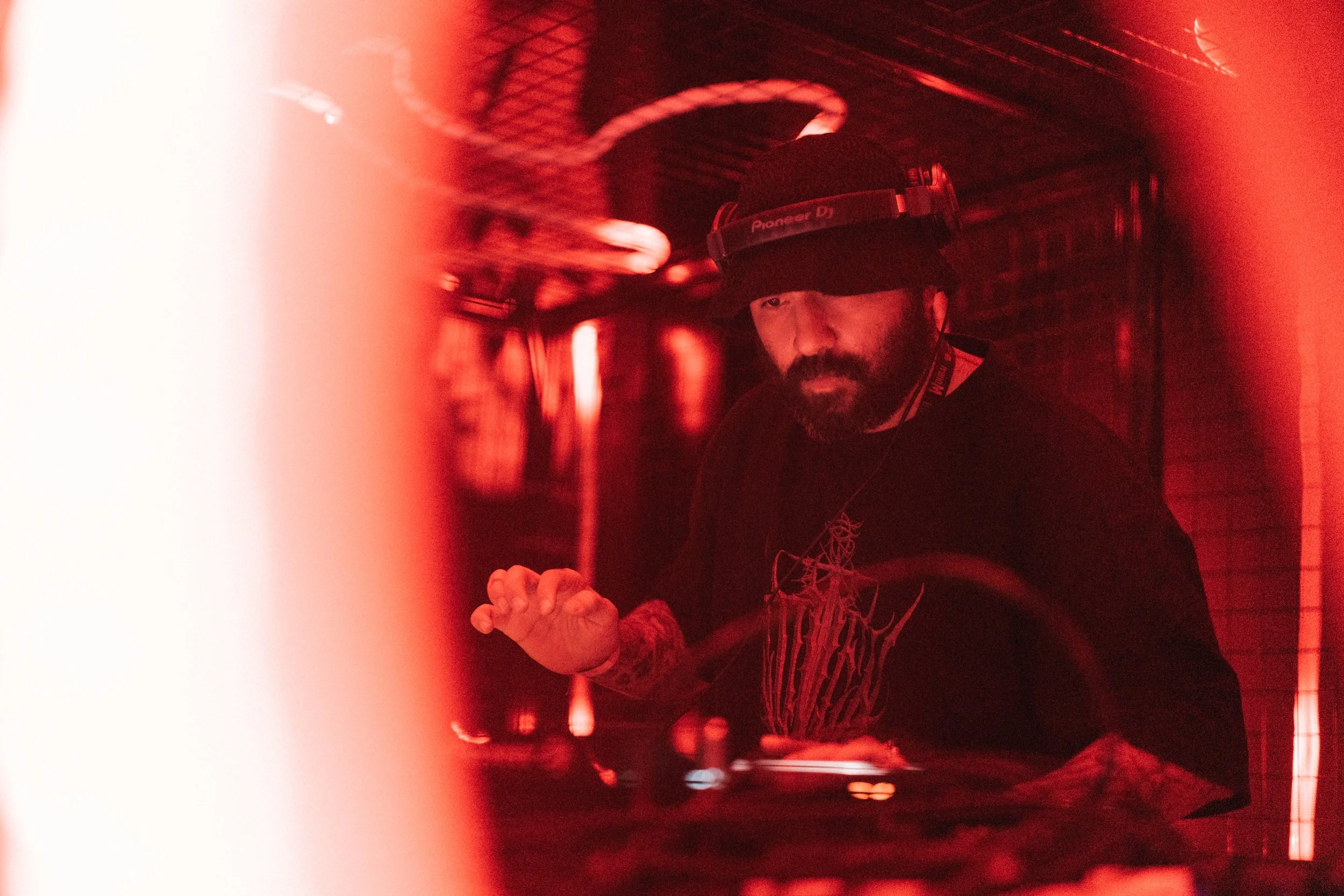 A DJ with a beard wearing a Pioneer DJ headband and a black shirt, playing music under red lighting in a dimly lit setting.