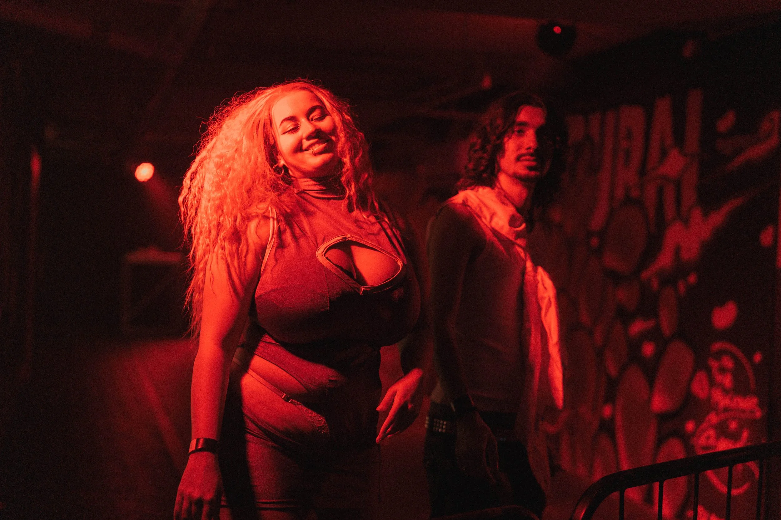 A woman with long, curly red hair smiling and a man with long dark hair standing in a dimly lit club with red lighting, with graffiti-style art on the wall in the background.