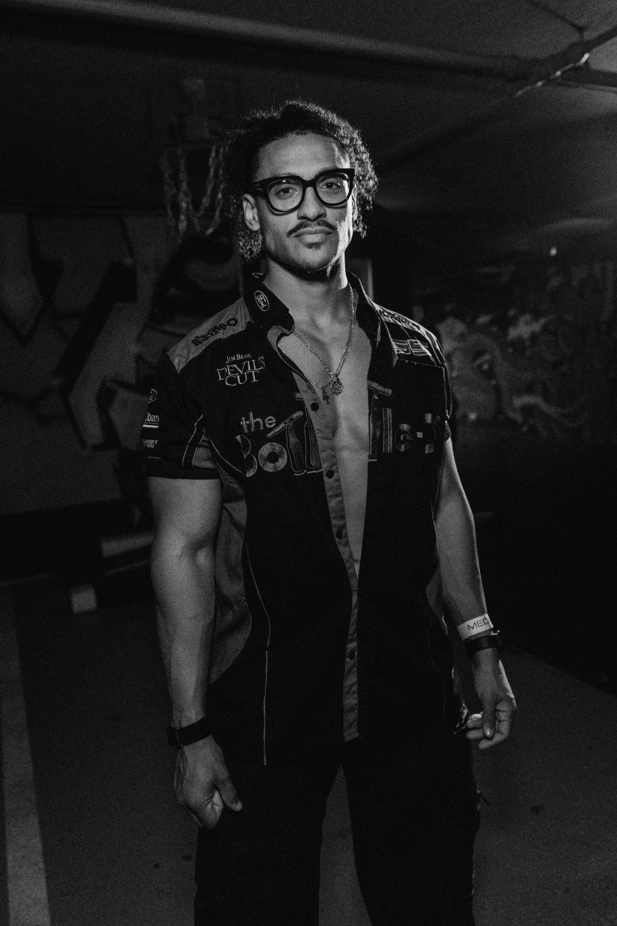 A young man with glasses and curly hair stands in a dimly lit room, wearing a partially unbuttoned racing shirt with various patches and logos. He has a necklace and wristband.