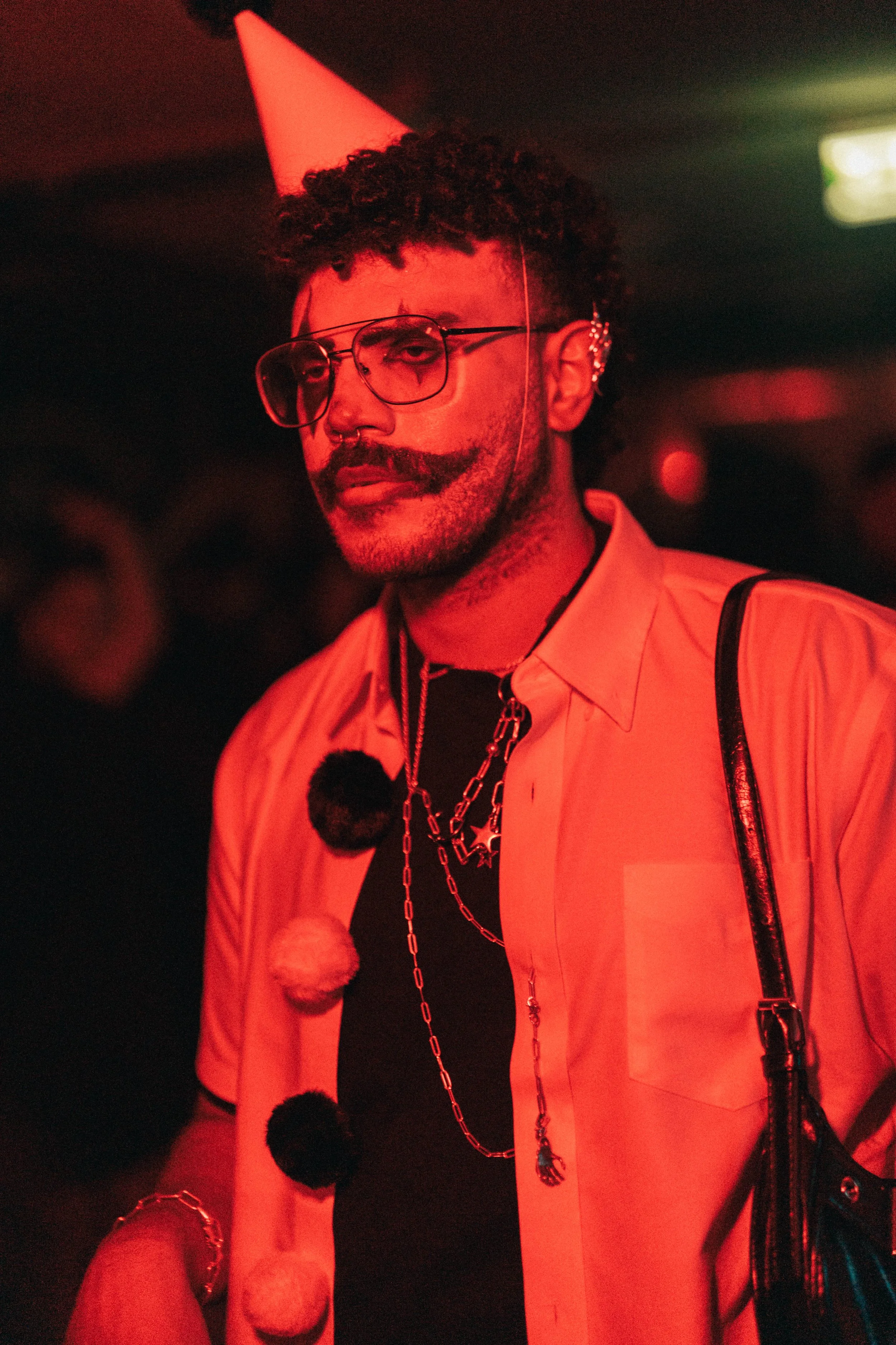 Person wearing a pink short-sleeved shirt, glasses, and a party hat, with colorful chain necklaces and pom-pom decorations, standing in a dimly lit environment.