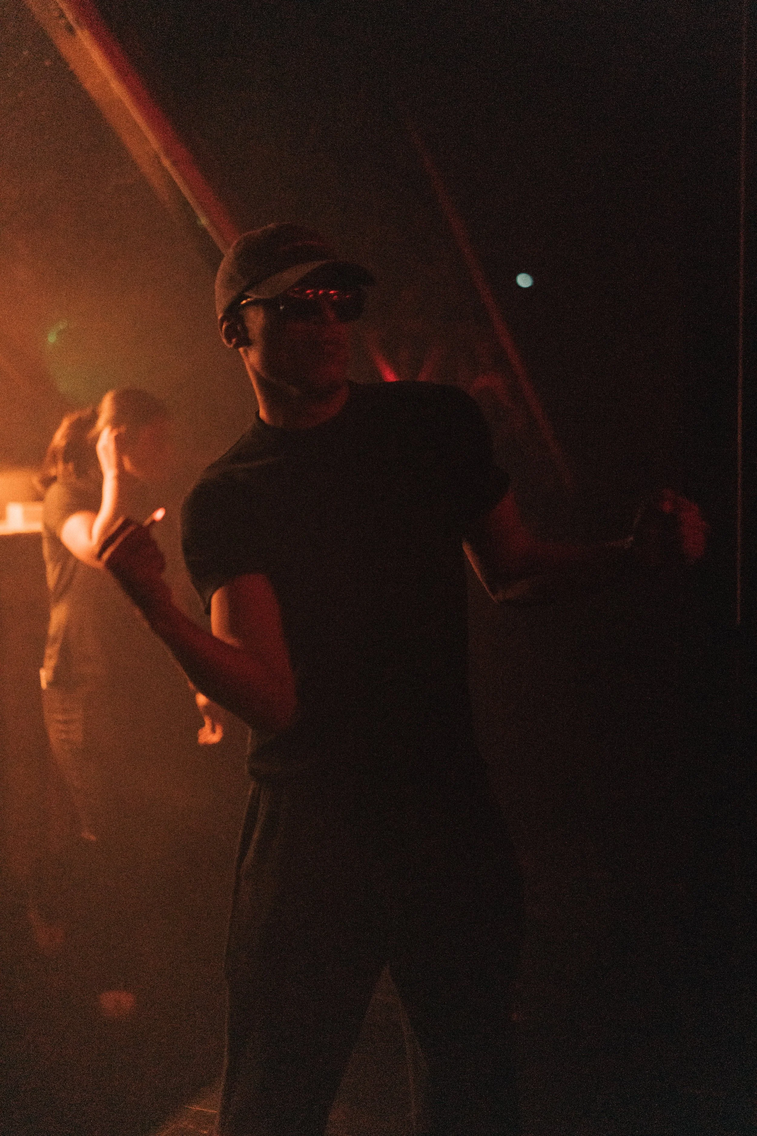 A person wearing a baseball cap and sunglasses in a dark, smoky environment, giving a thumbs-up gesture.