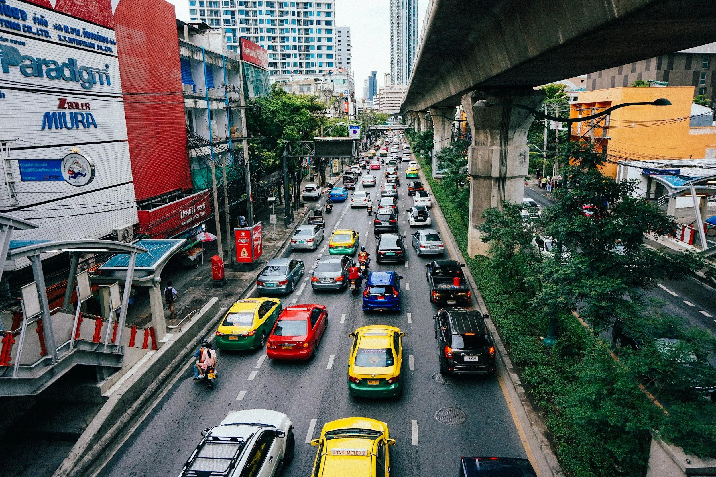 A busy city street filled with cars, including taxis, and a motorcycle, with an elevated train track overhead, surrounded by tall buildings and greenery.