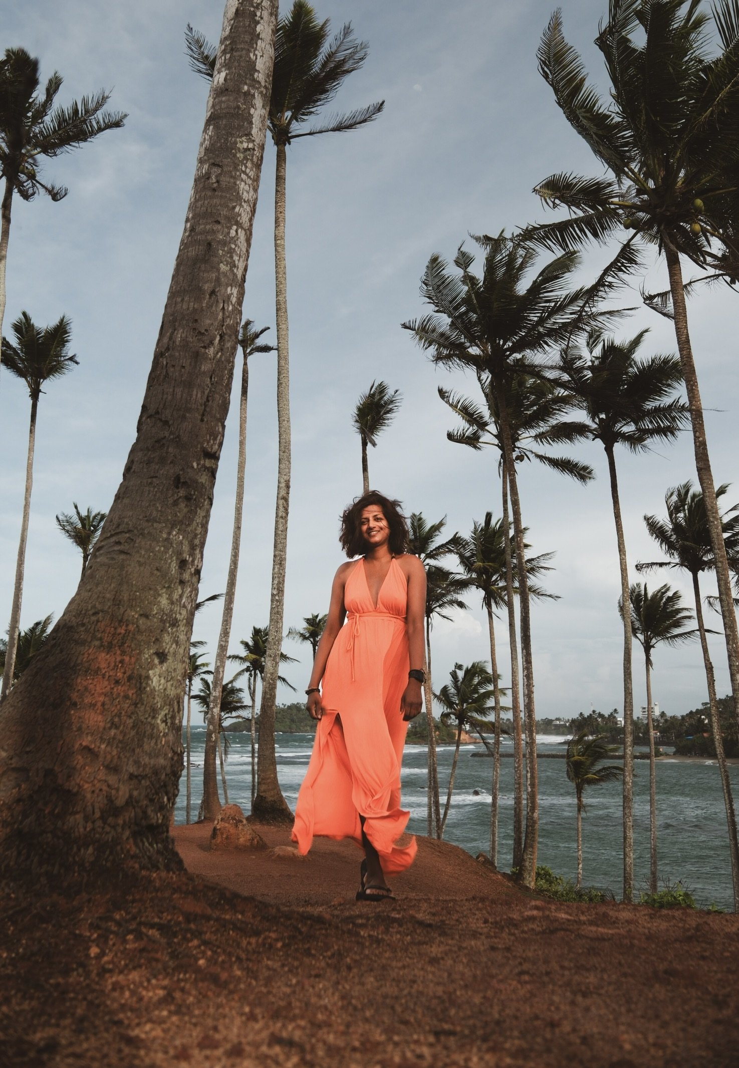 A woman in an orange dress walking along a tropical beach with tall palm trees and water in the background.