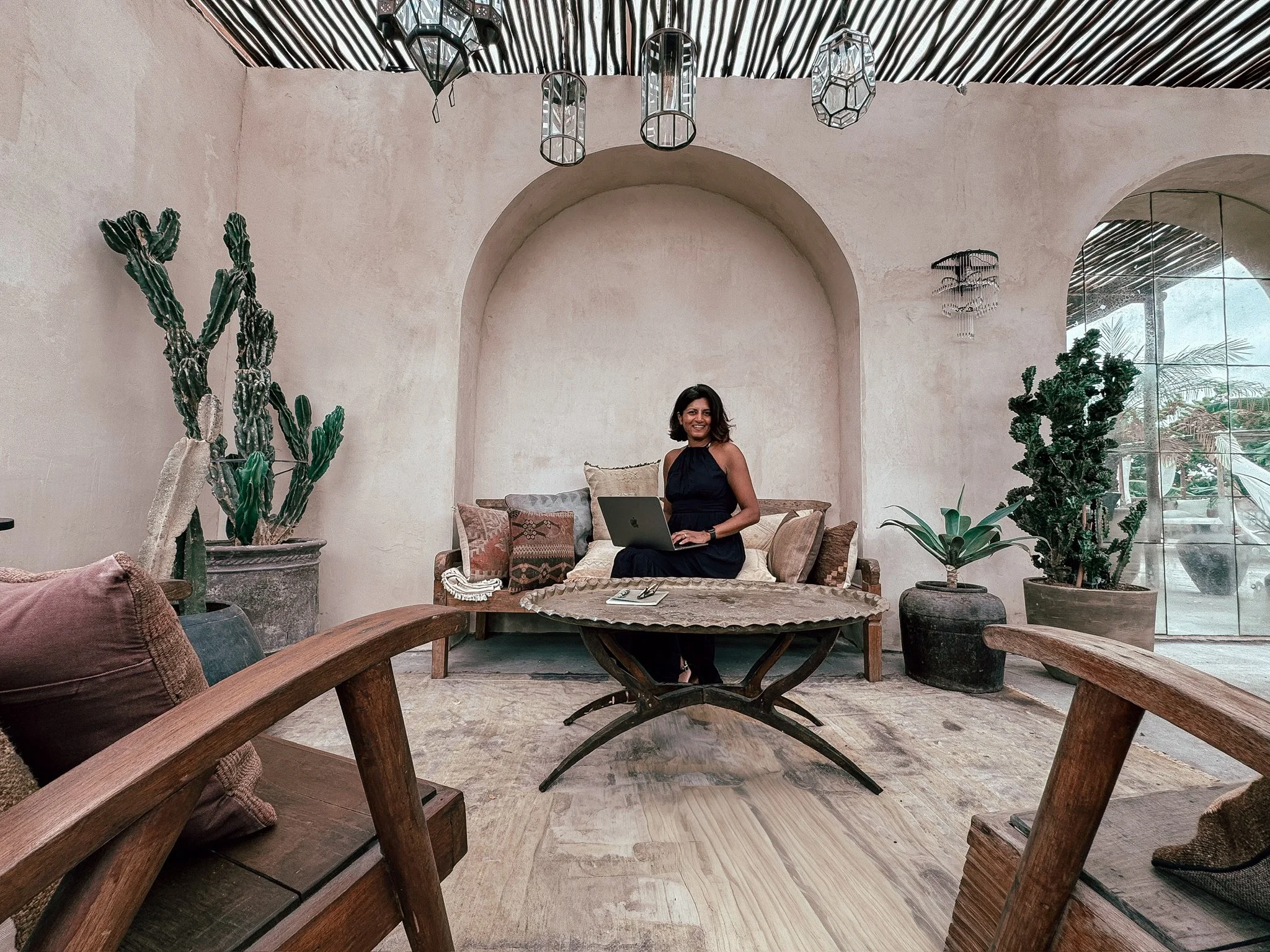 A woman sitting on a beige sofa with pillows, using a laptop in a stylish indoor space with large potted cacti and plants, rustic decor, a wooden coffee table, and a glass door showing an outdoor area.