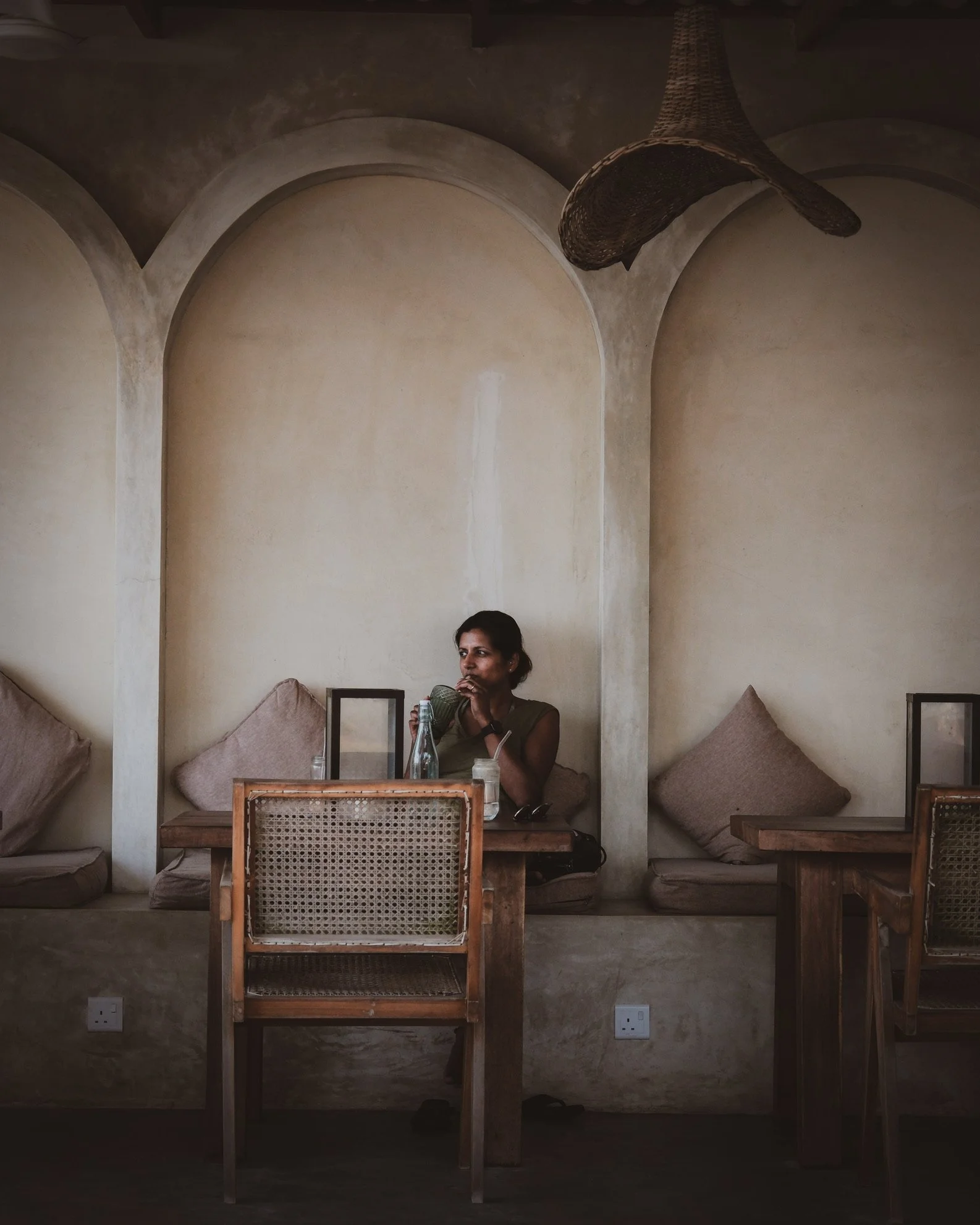 A woman sitting at a wooden table in a dimly lit, rustic cafe with beige walls and arched alcoves, sipping from a glass bottle, with a glass of water and a face mask on the table.