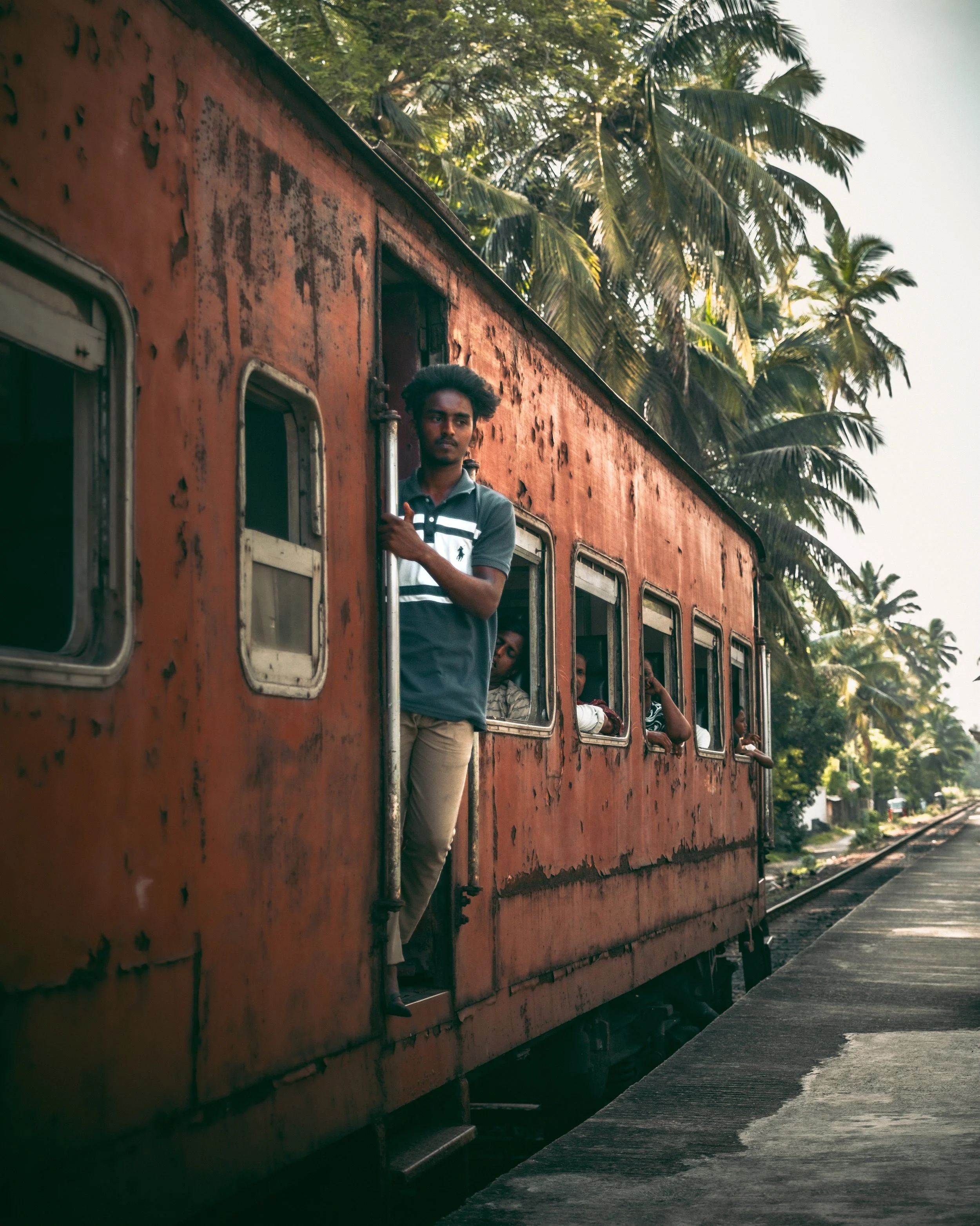 Man standing at the door of an old, rusted red train on a railway track surrounded by palm trees.