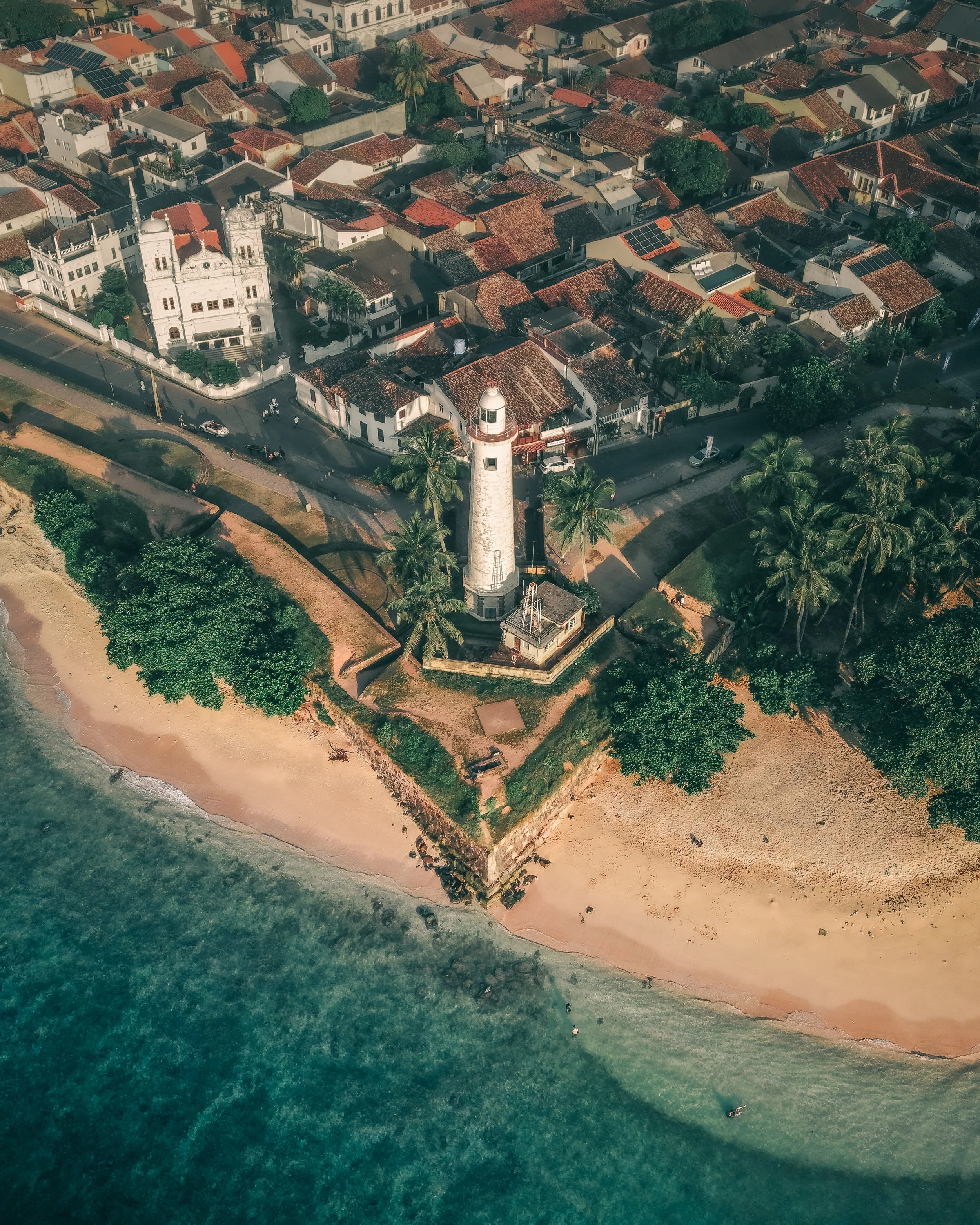 An aerial view of a coastal town showing a white lighthouse on a small peninsula, surrounded by palm trees, residential houses with red-tiled roofs, and a sandy beach along the shoreline, with clear ocean water.