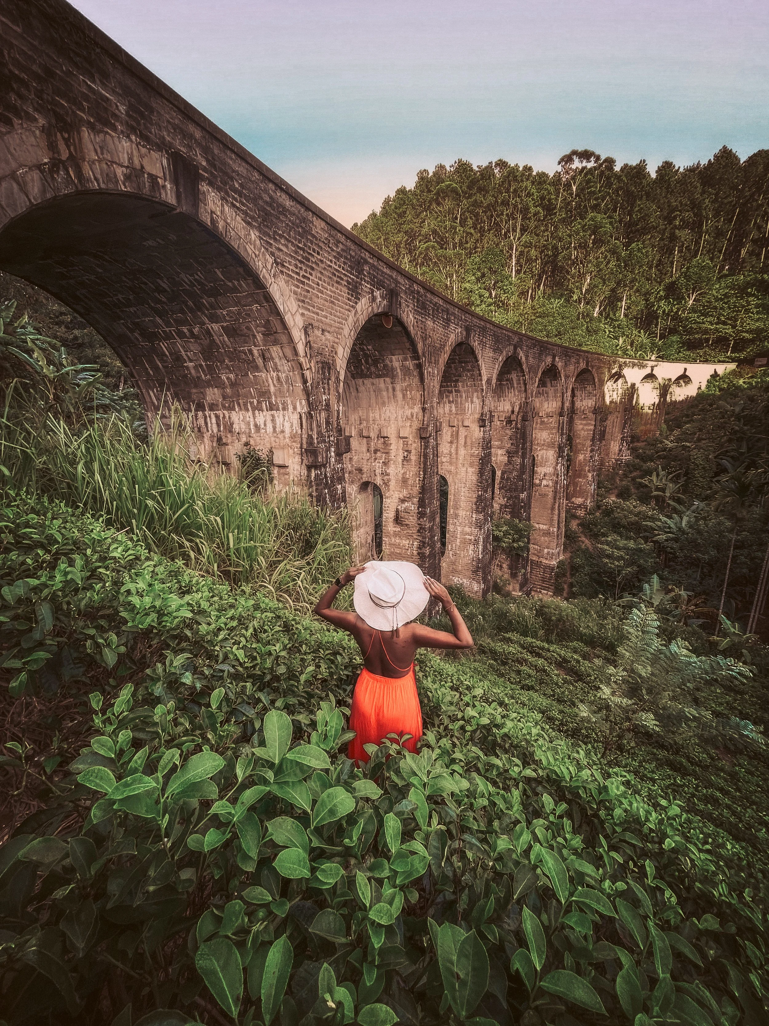 A woman in an orange dress and white hat standing among green plants near an old stone arch bridge in a lush, hilly landscape.